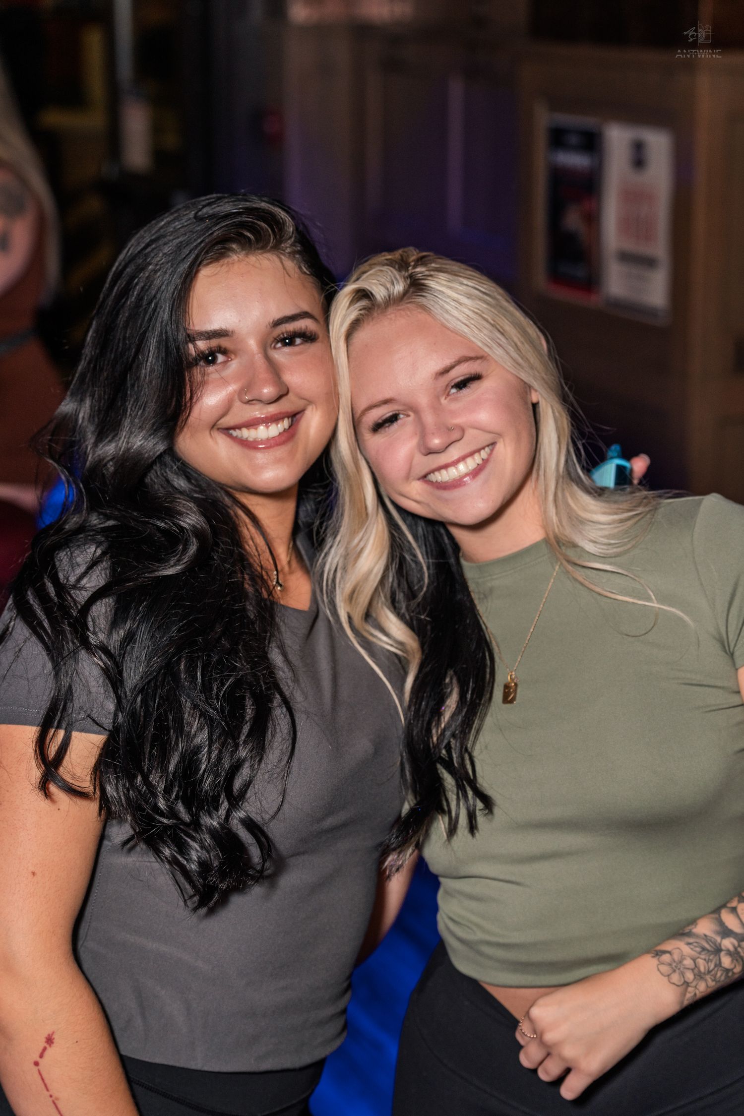 Two smiling women pose together at a nightclub, one with dark hair in gray top and one blonde in green top.