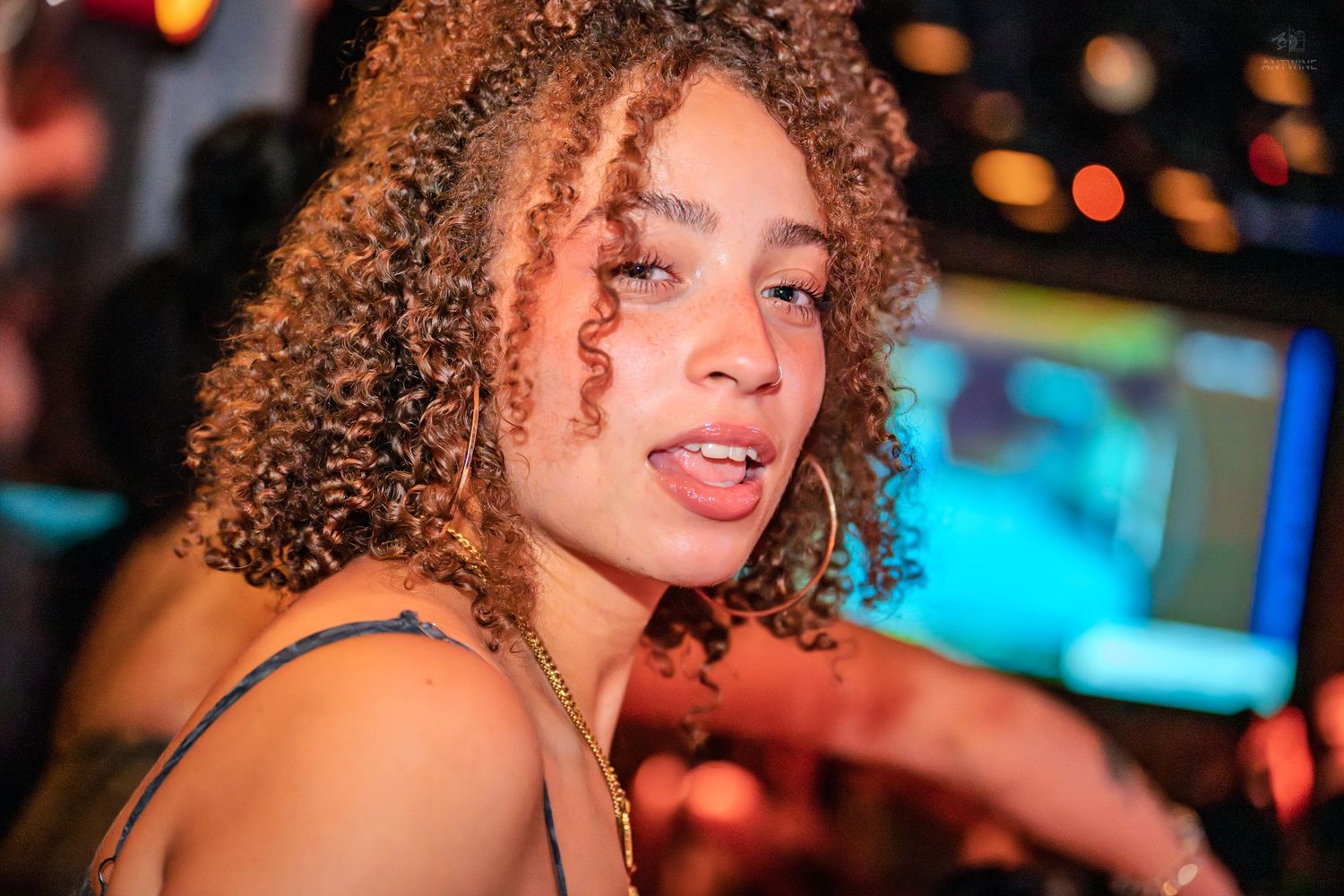 Young woman with curly hair smiling at a nightclub event with colorful lights in the background.