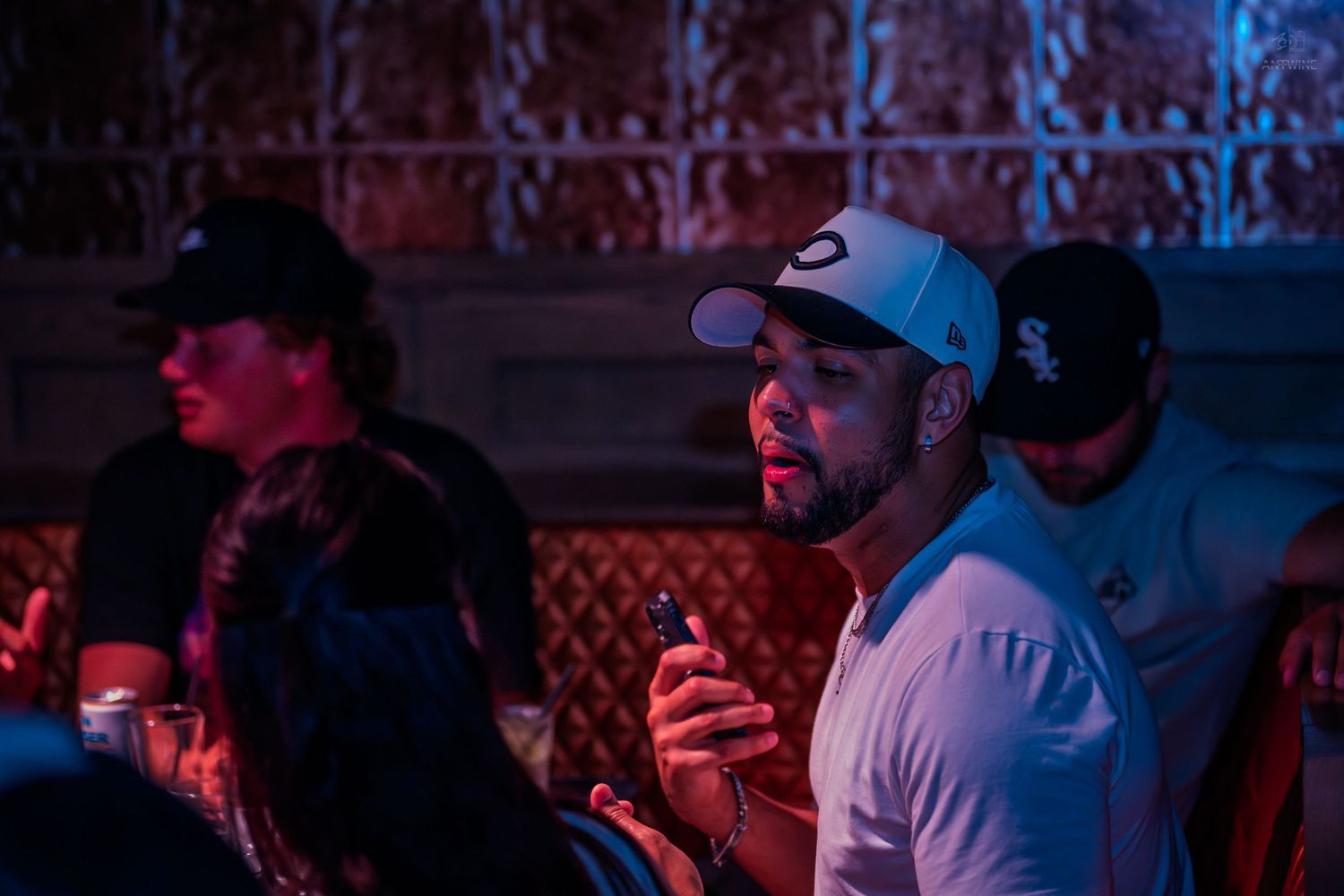 Man in white shirt and baseball cap holding phone at dimly lit nightclub event.