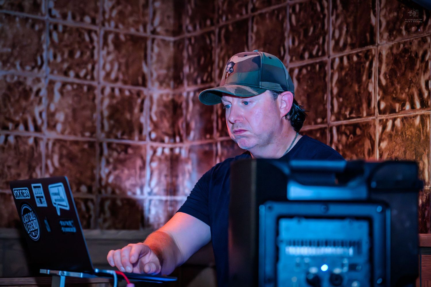 Man in a camo cap working on a laptop at a music event or DJ setup.