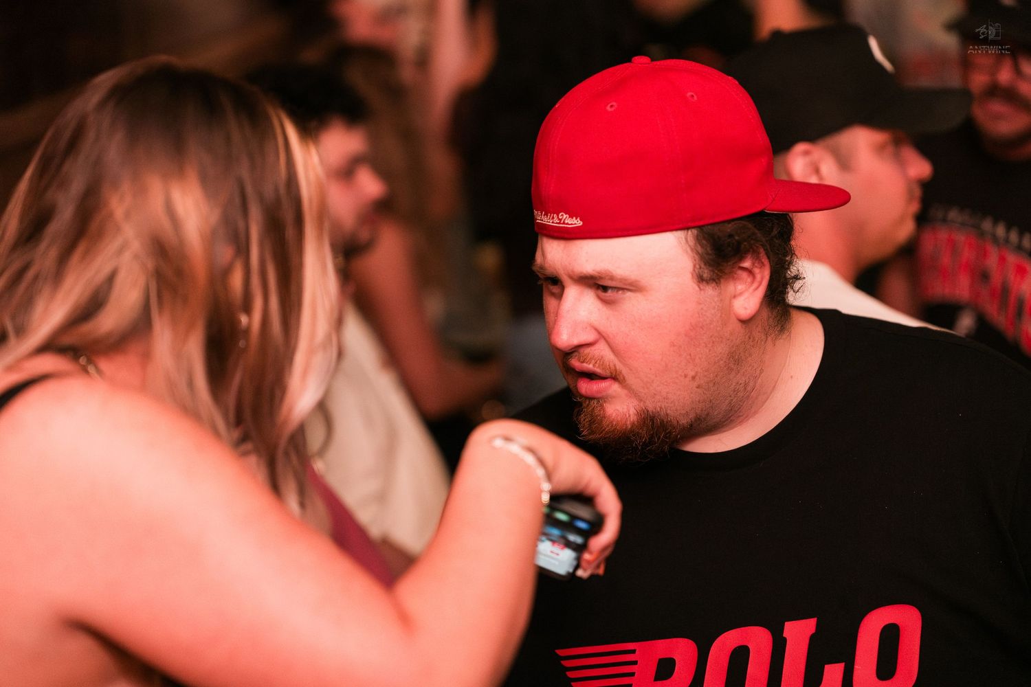 Man in red cap and POLO shirt holds drink while chatting with woman at a crowded nightclub event.