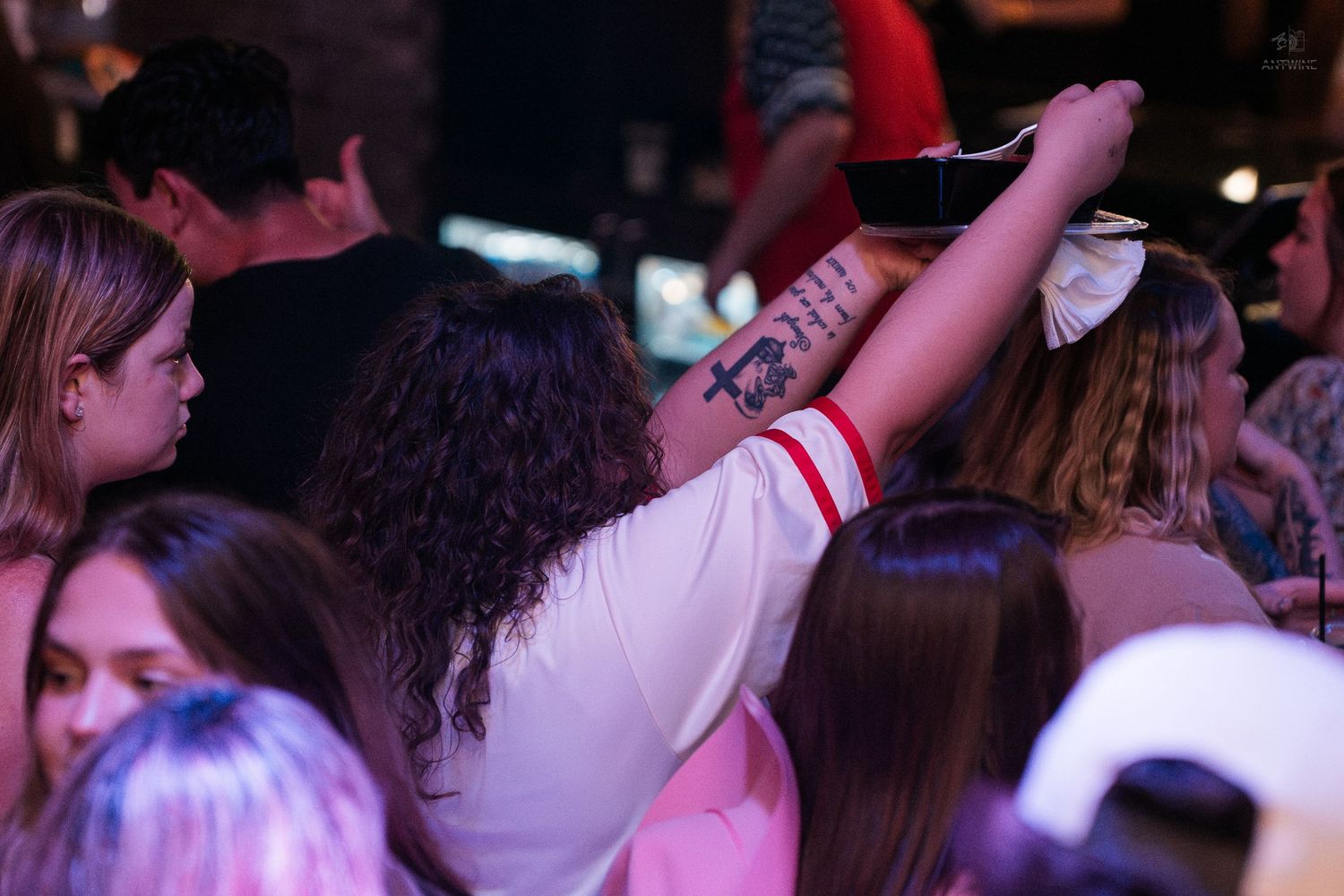 Woman with tattoos raises a drink while dancing in a crowded nightclub with friends.