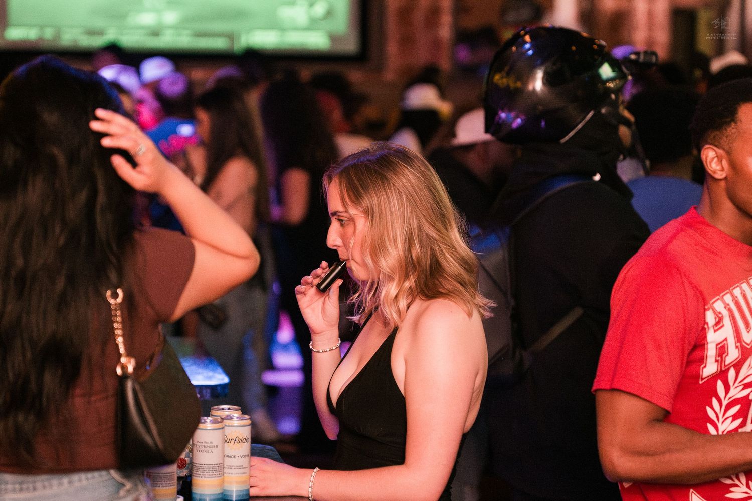 Young woman sips a drink while standing in a packed nightclub with colorful lighting effects.