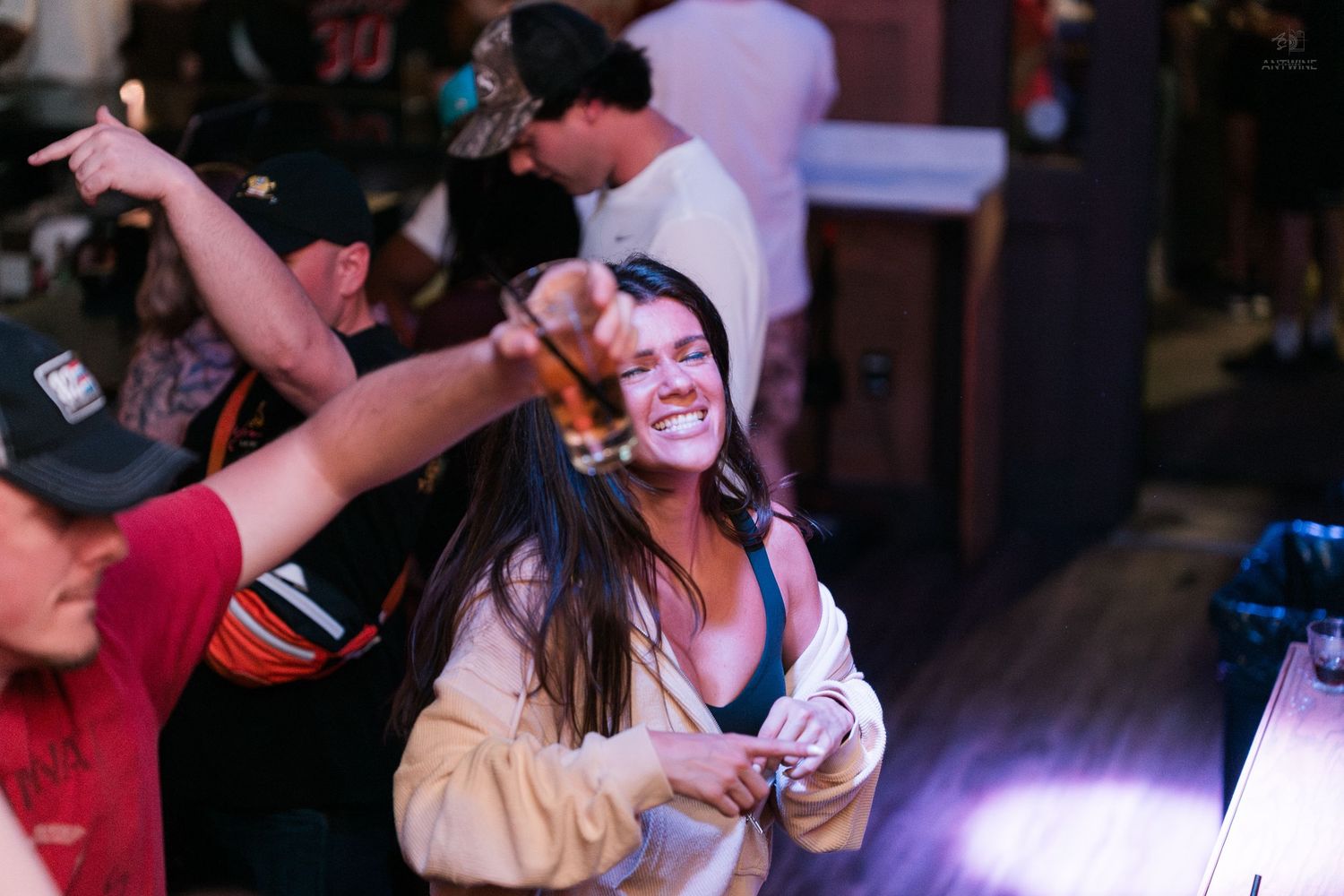 A smiling young woman with long dark hair dances joyfully while holding a drink at a crowded bar.