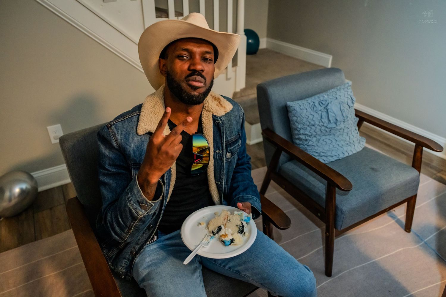 Man in cowboy hat and denim jacket sitting in a chair holding a plate of birthday cake.