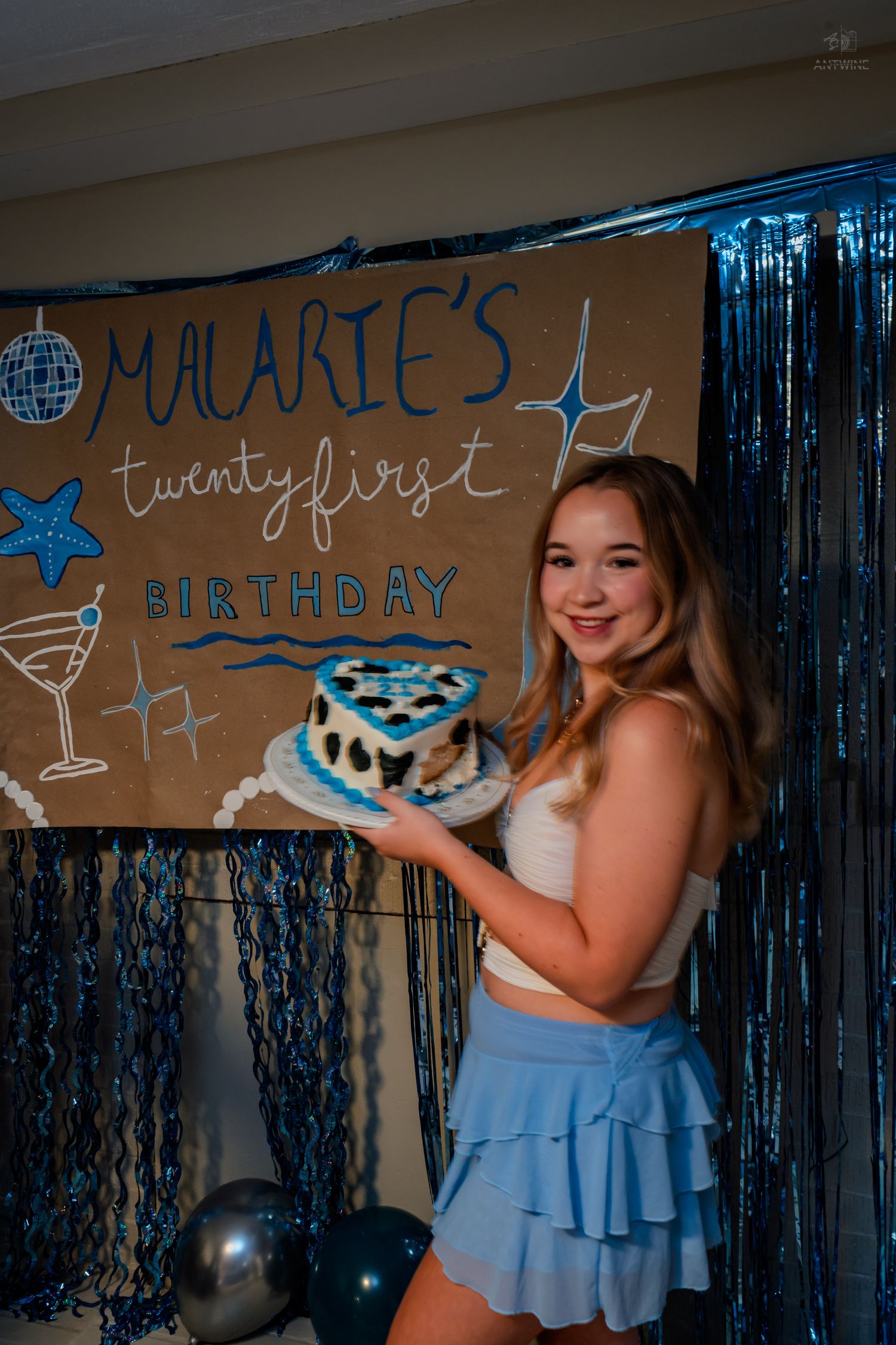 Smiling birthday girl holds blue heart-shaped cake at Malarie's twenty-first birthday party with blue tinsel curtains.