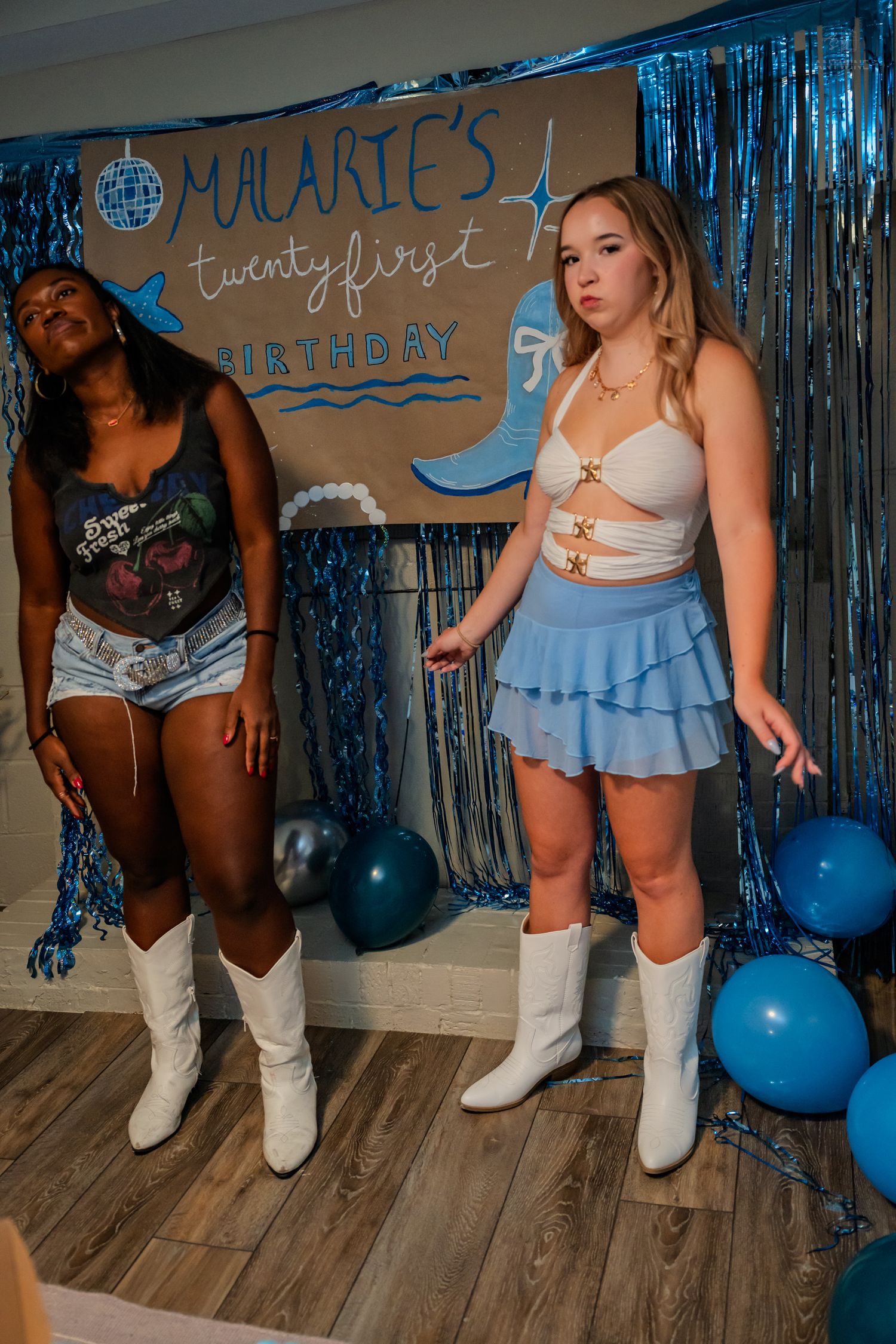 Two women in coordinated outfits and white cowboy boots pose at a 21st birthday party.