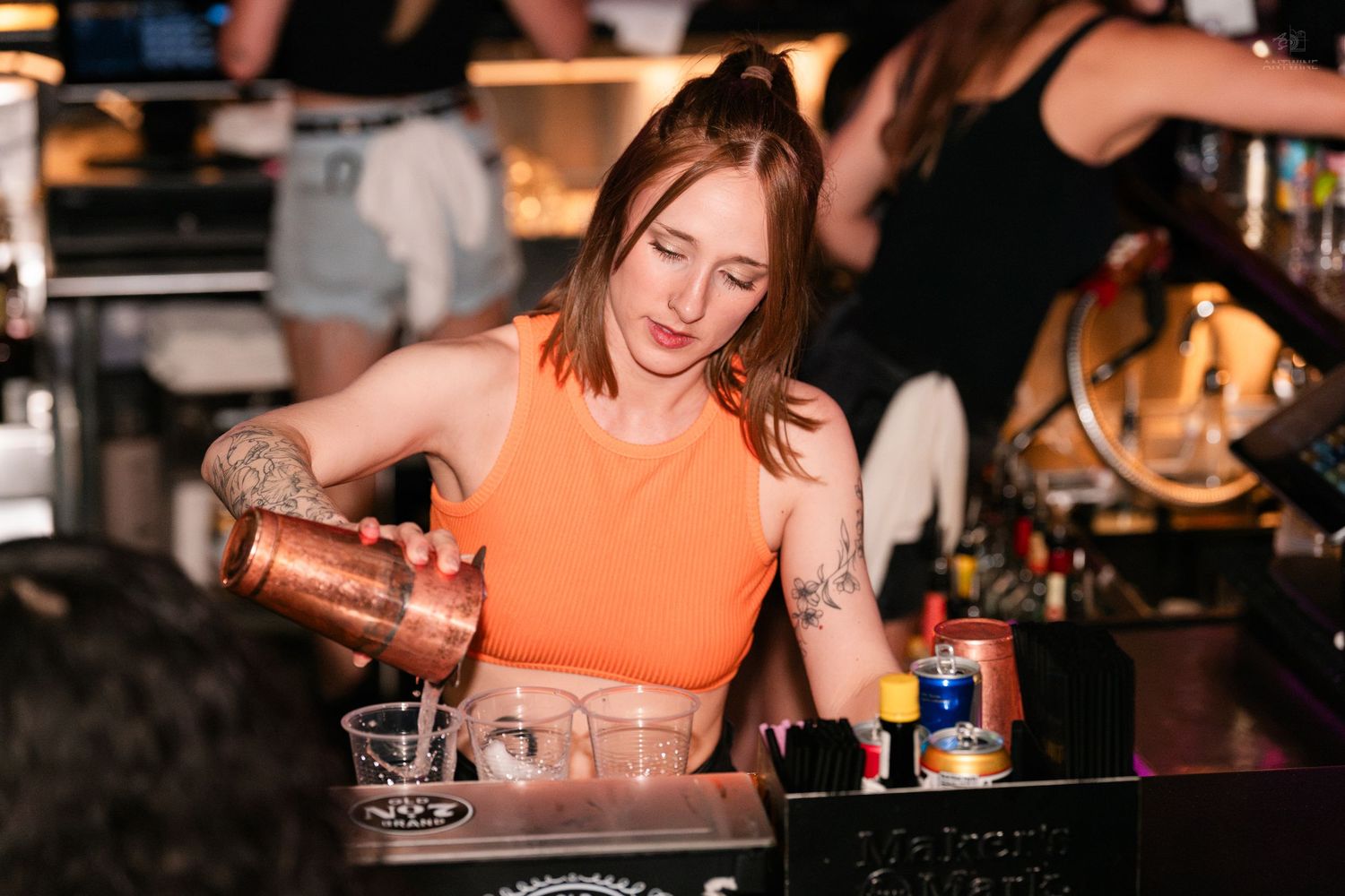 Tattooed female bartender in orange top pours drinks at a nightclub bar.