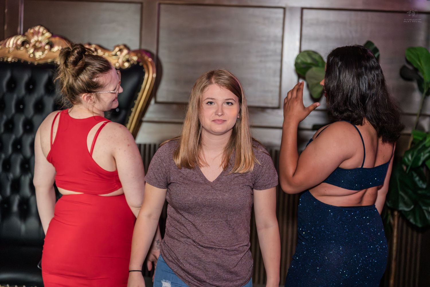 Three women at a nightclub, one in mauve top looks back while two others face away near ornate chair.
