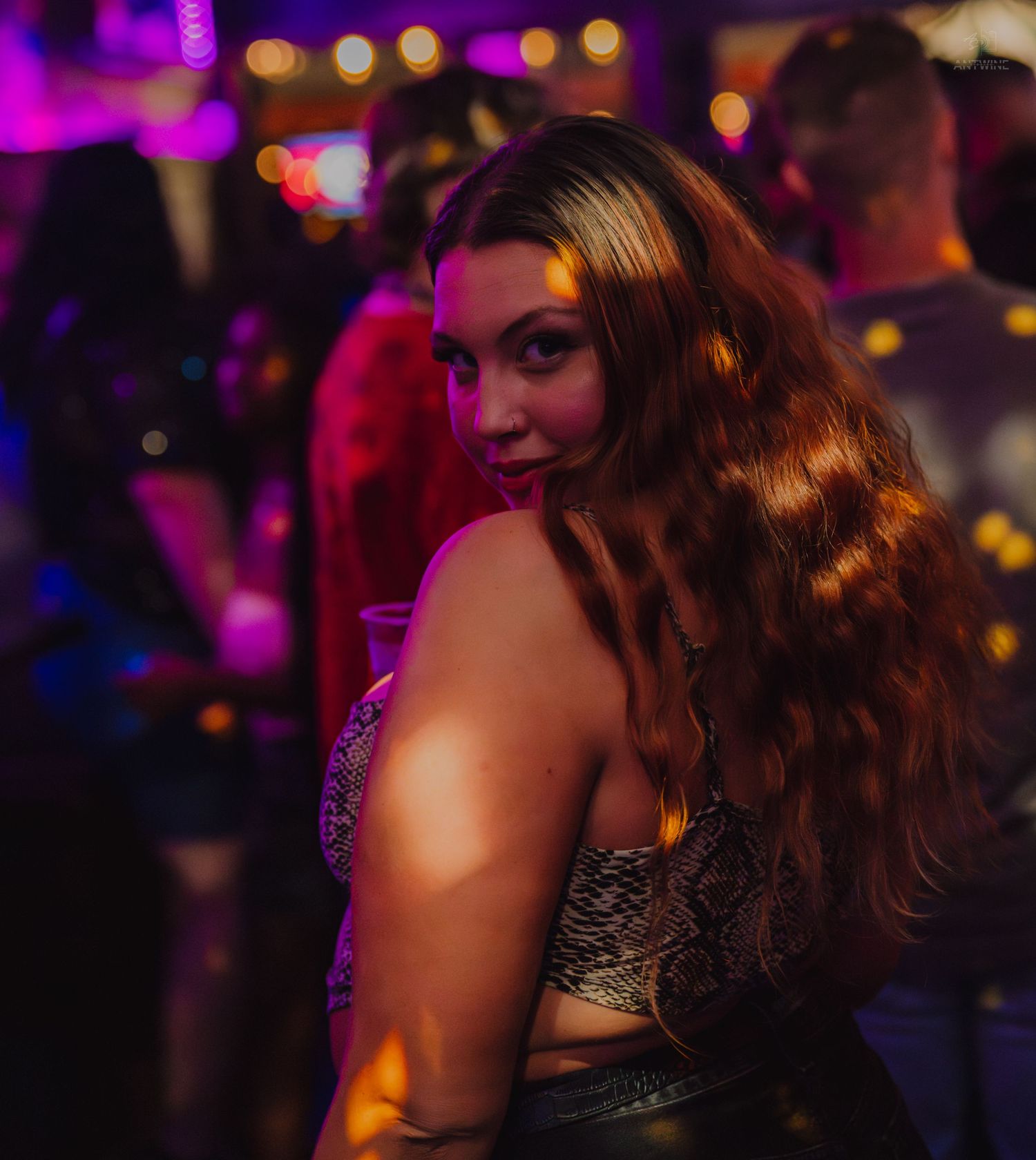 Young woman with long wavy hair posing at a vibrant nightclub with purple and pink neon lights.