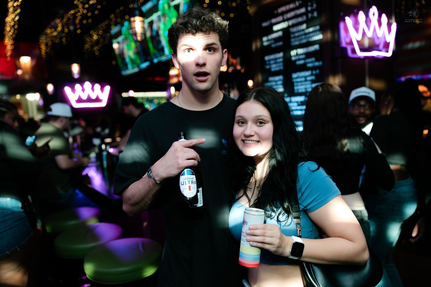 Young couple smiling and holding drinks at a colorful nightclub venue.