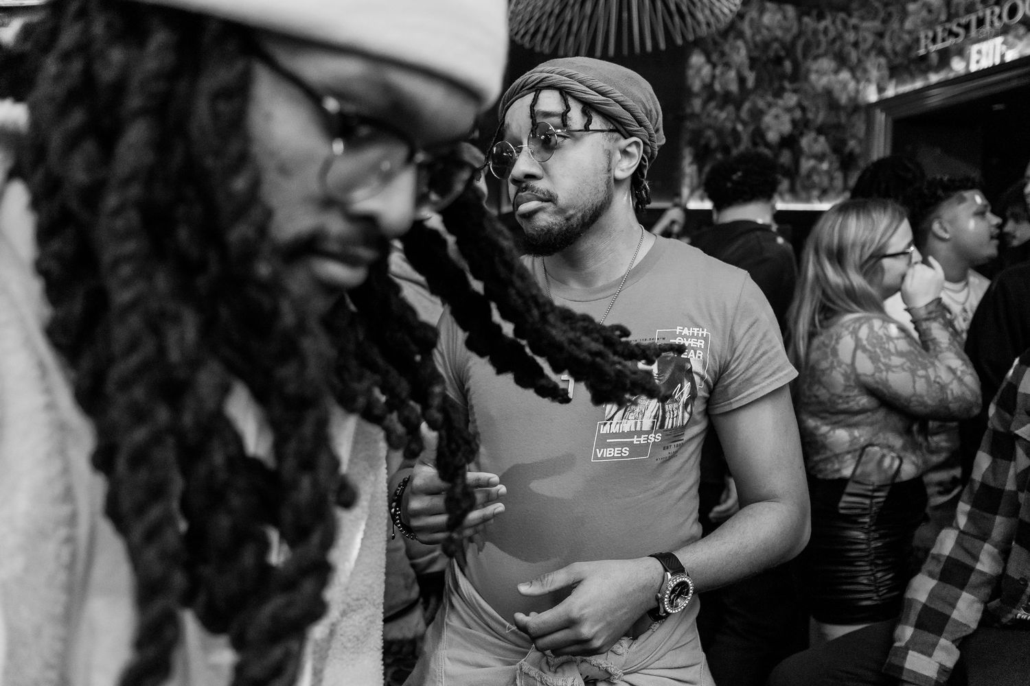 Two men with dreadlocks standing in a crowded venue, one wearing reflective sunglasses in black and white photo.
