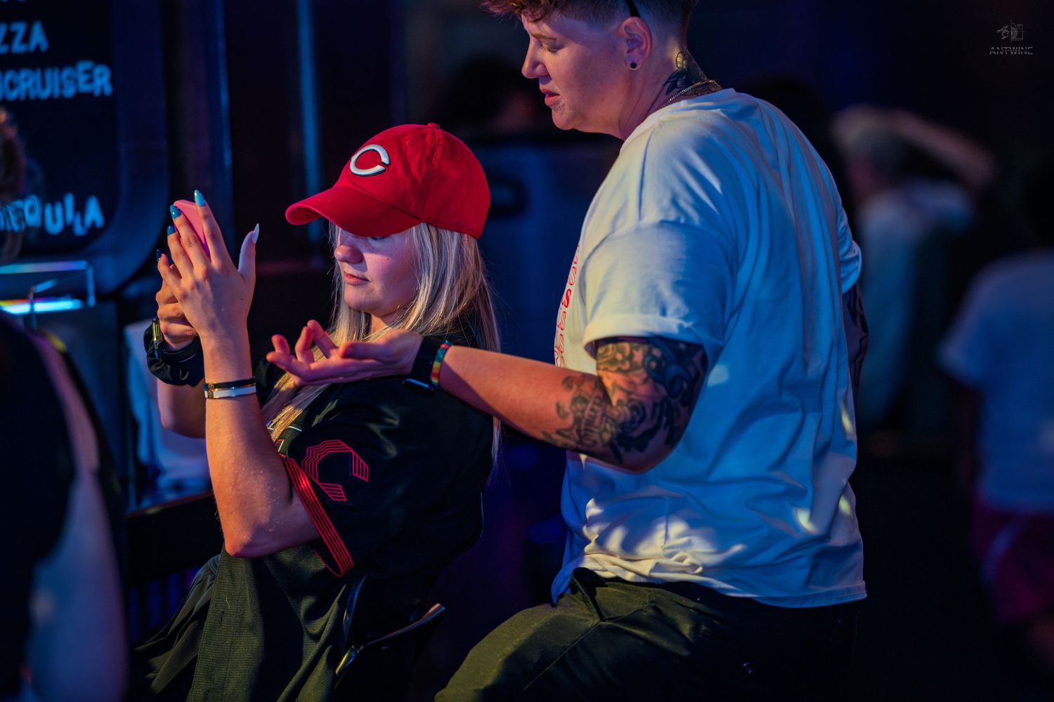 Woman in red cap clapping hands at a nightclub event with tattooed person nearby.