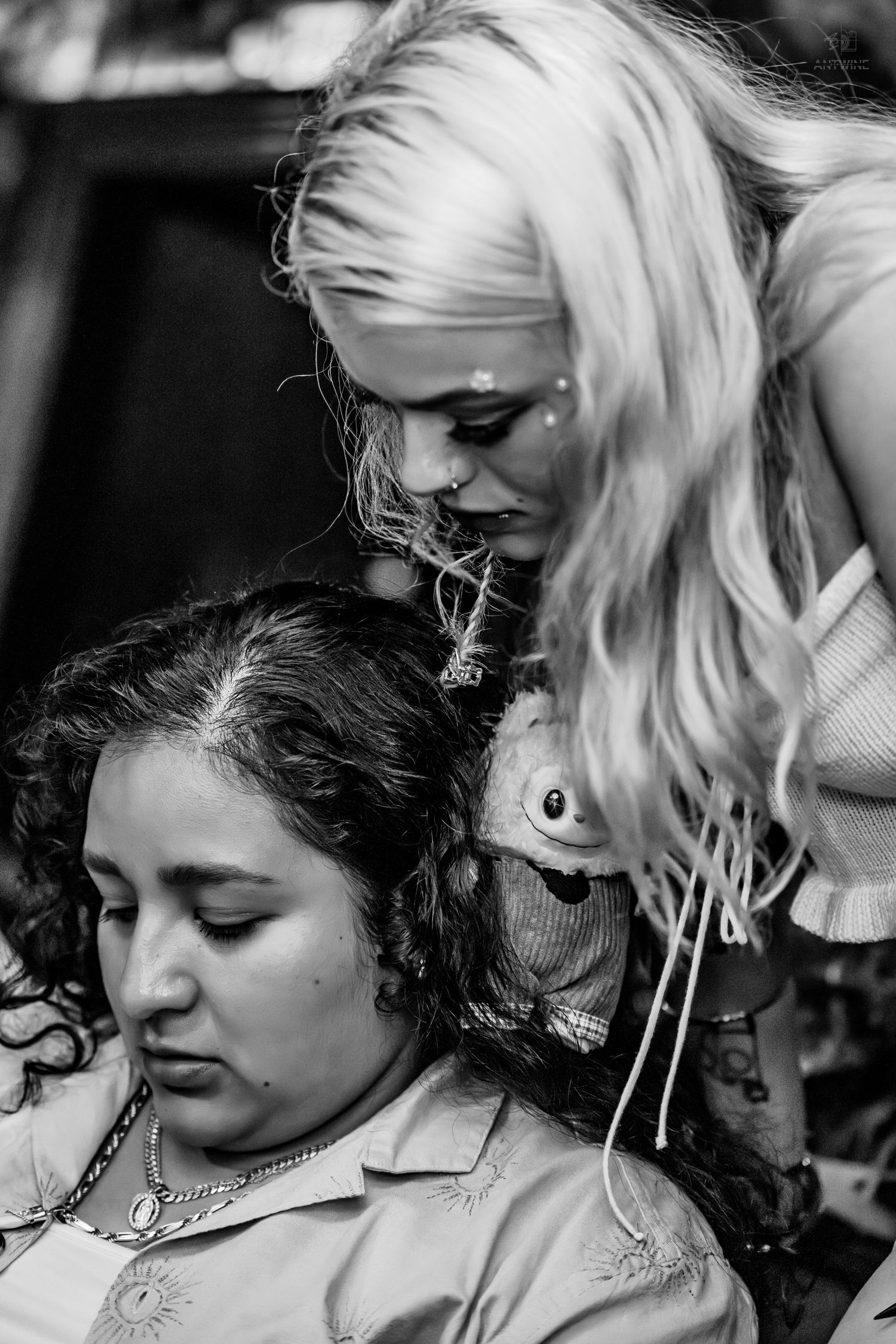 Black and white photo of blonde woman with face gems leaning over dark-haired woman at festival event.