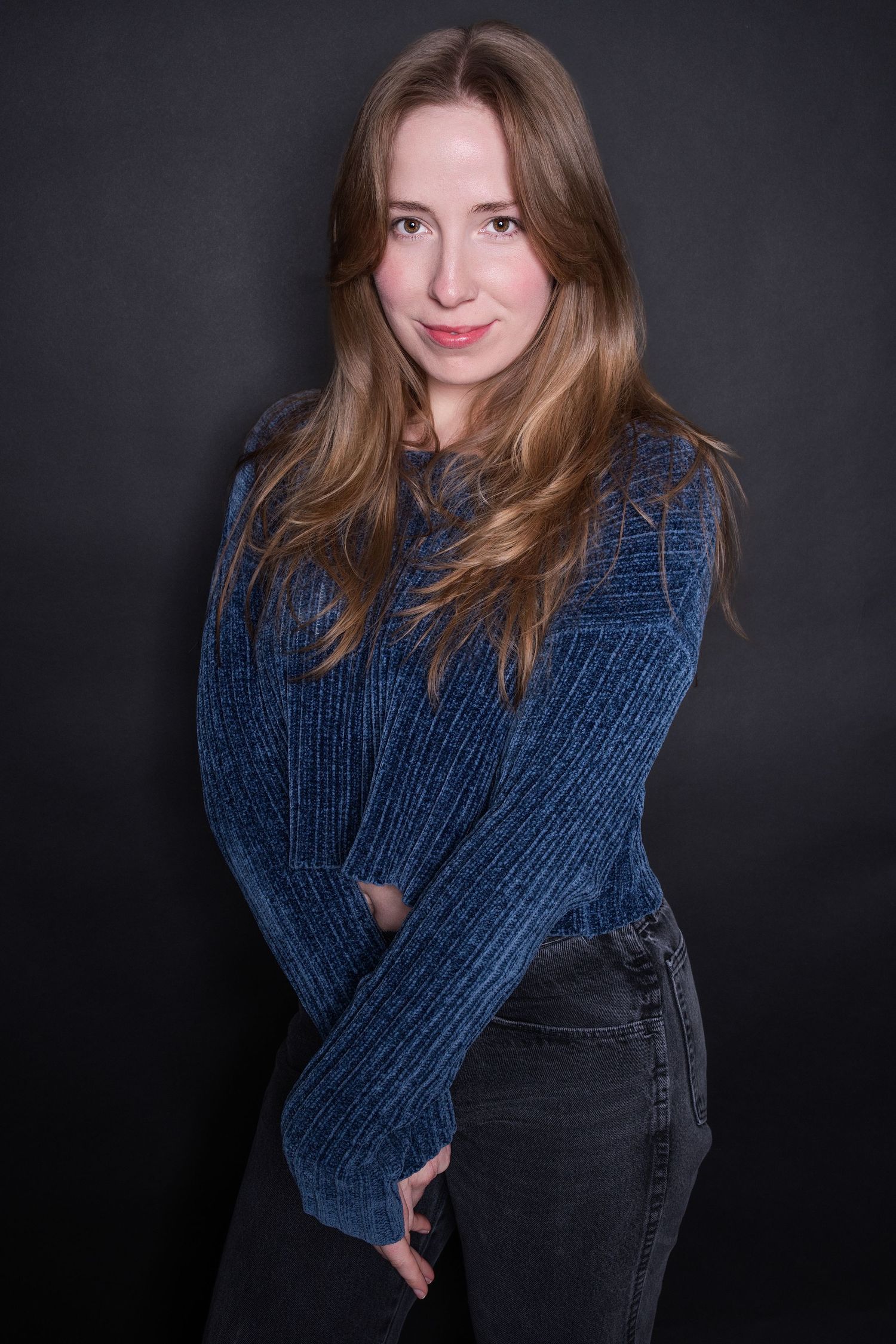 Young woman in a blue knit sweater and jeans poses for a studio portrait against dark background.