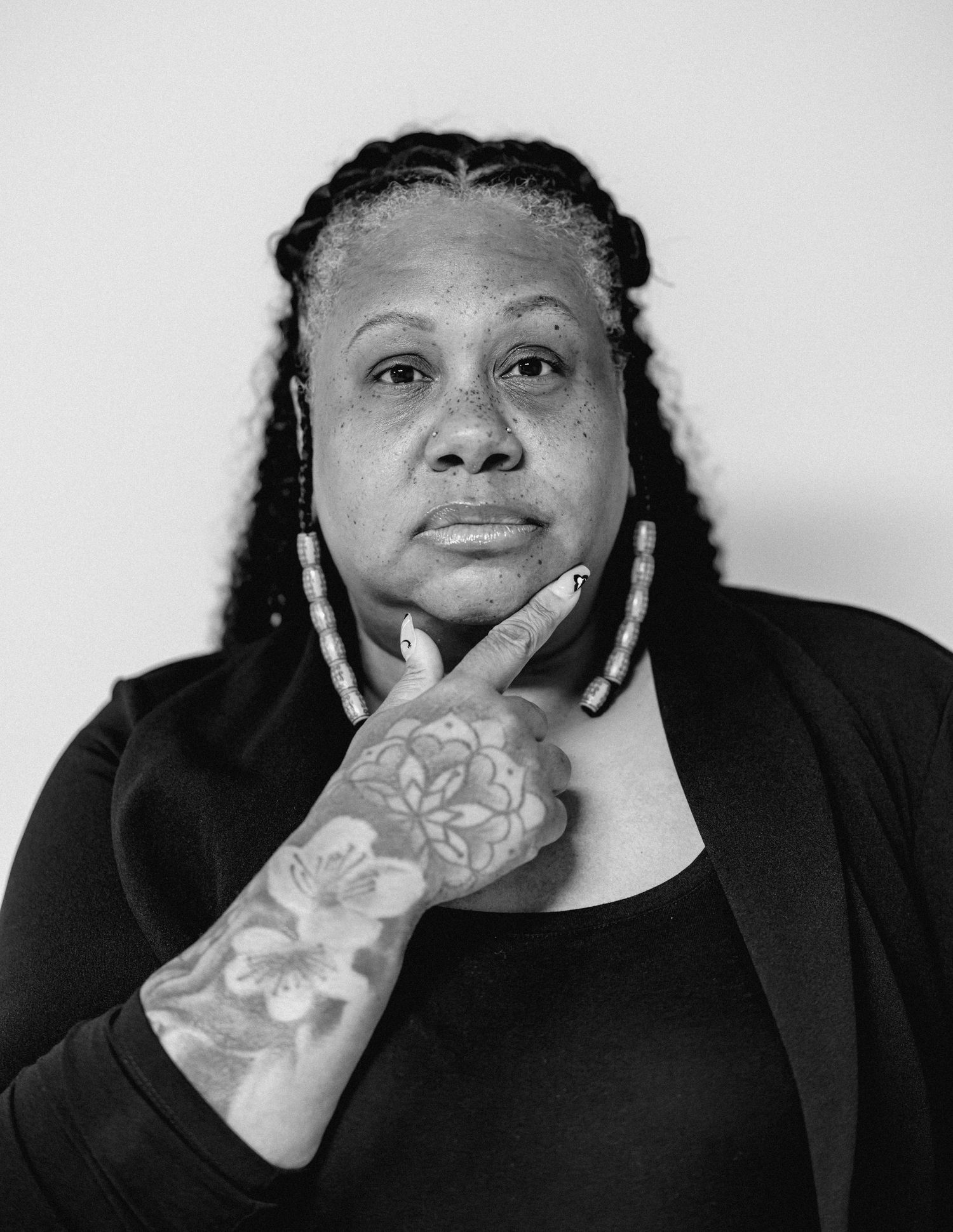 Black and white portrait of a woman with braided hair and floral hand tattoo resting hand on chin.
