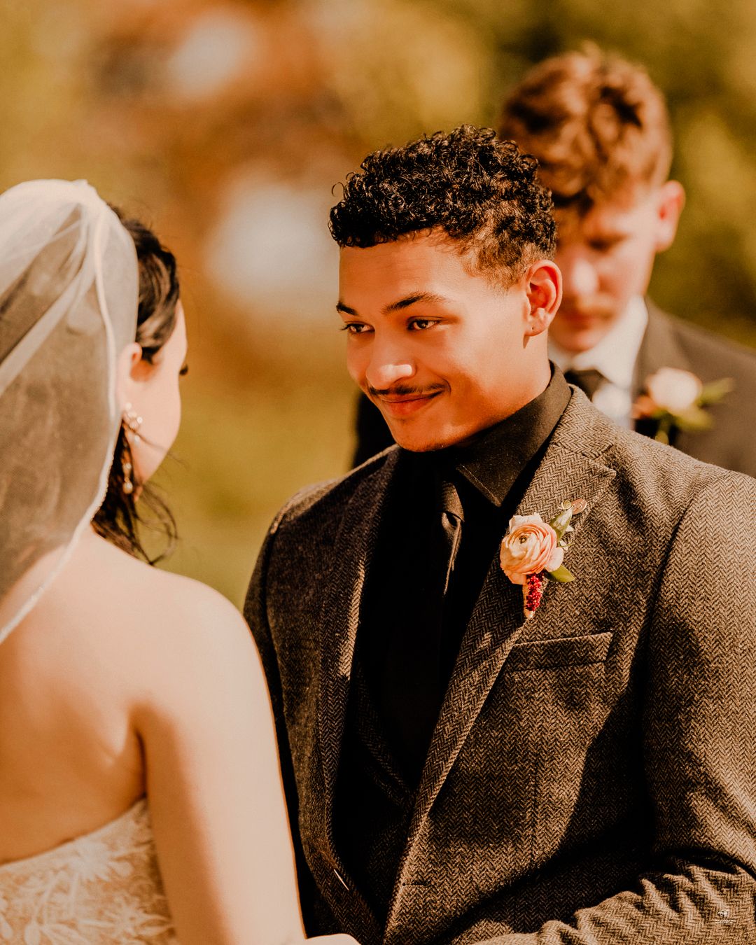 Groom in grey tweed suit with floral boutonniere smiling at bride during outdoor wedding ceremony.