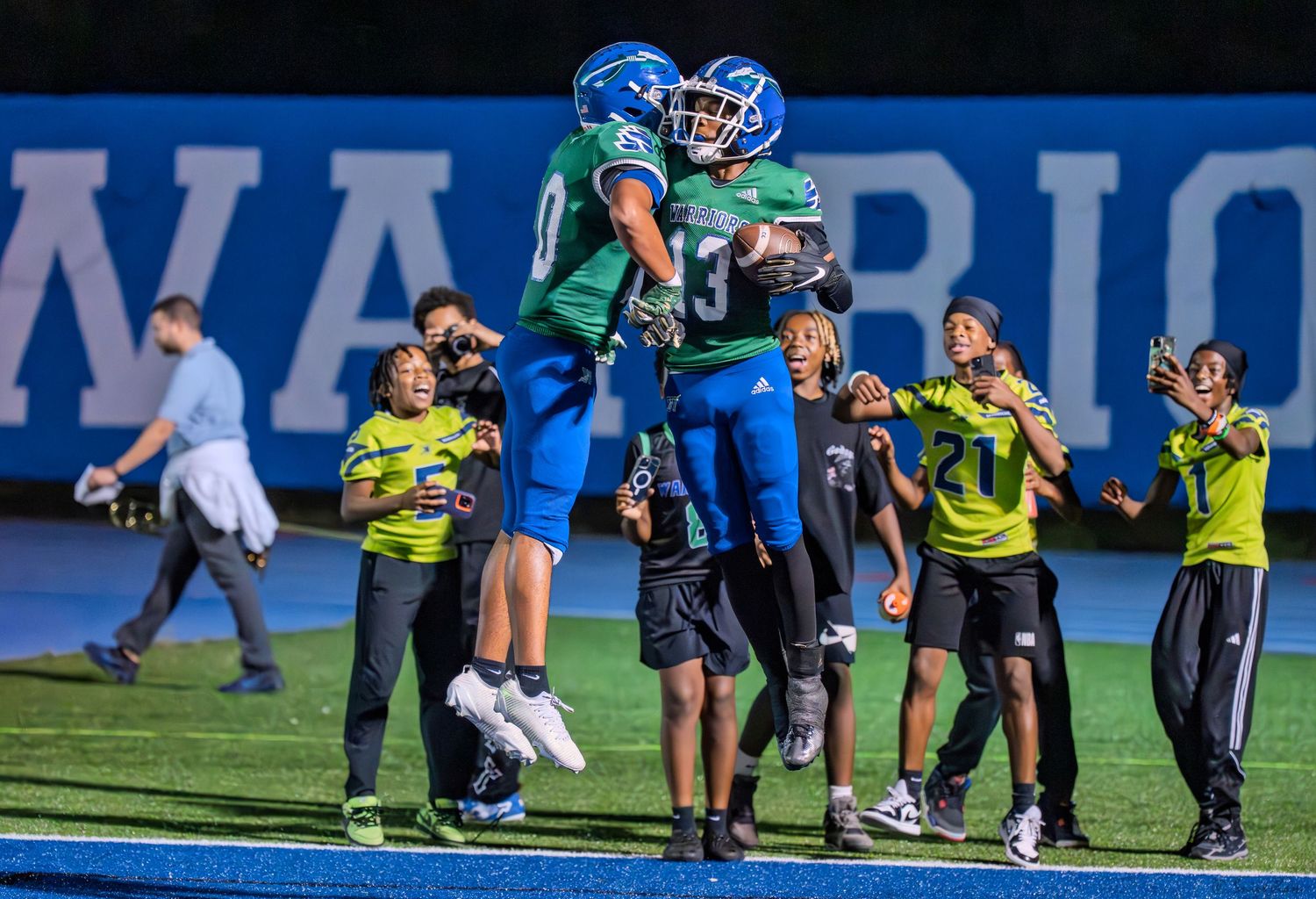 Two high school football players in green and blue uniforms celebrate with a chest bump after a score.