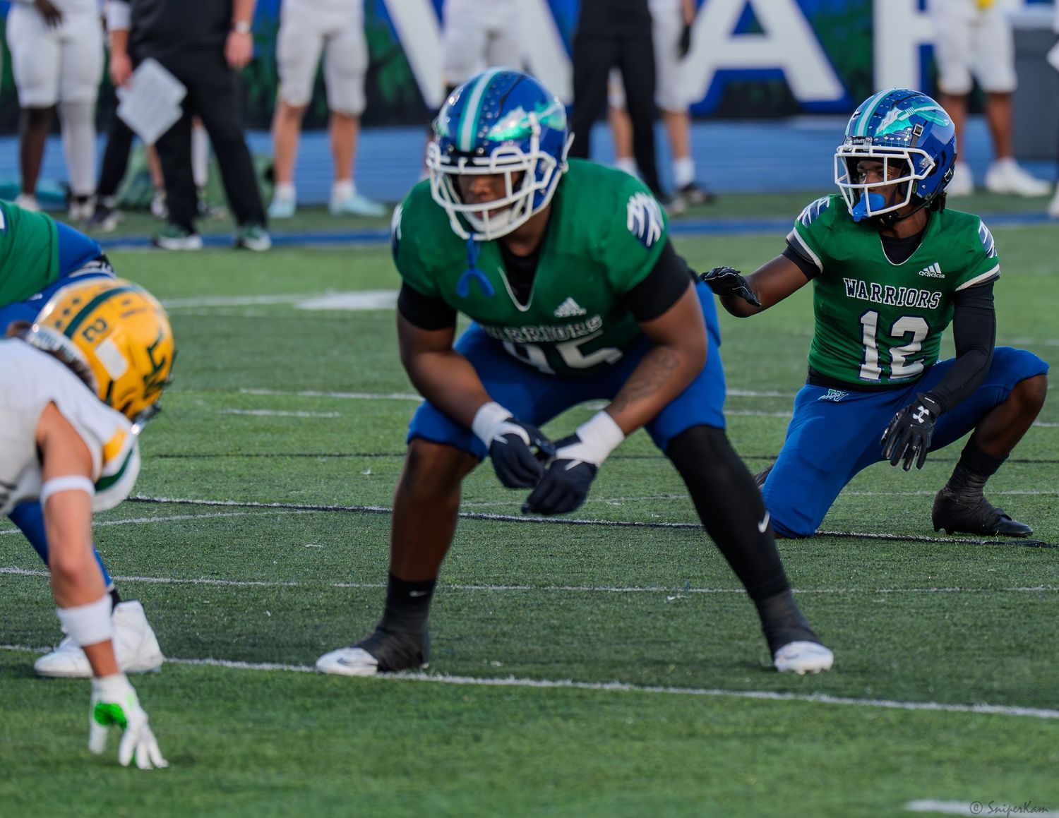 Two football players in teal uniforms crouch in defensive positions on the field during a high school football game.