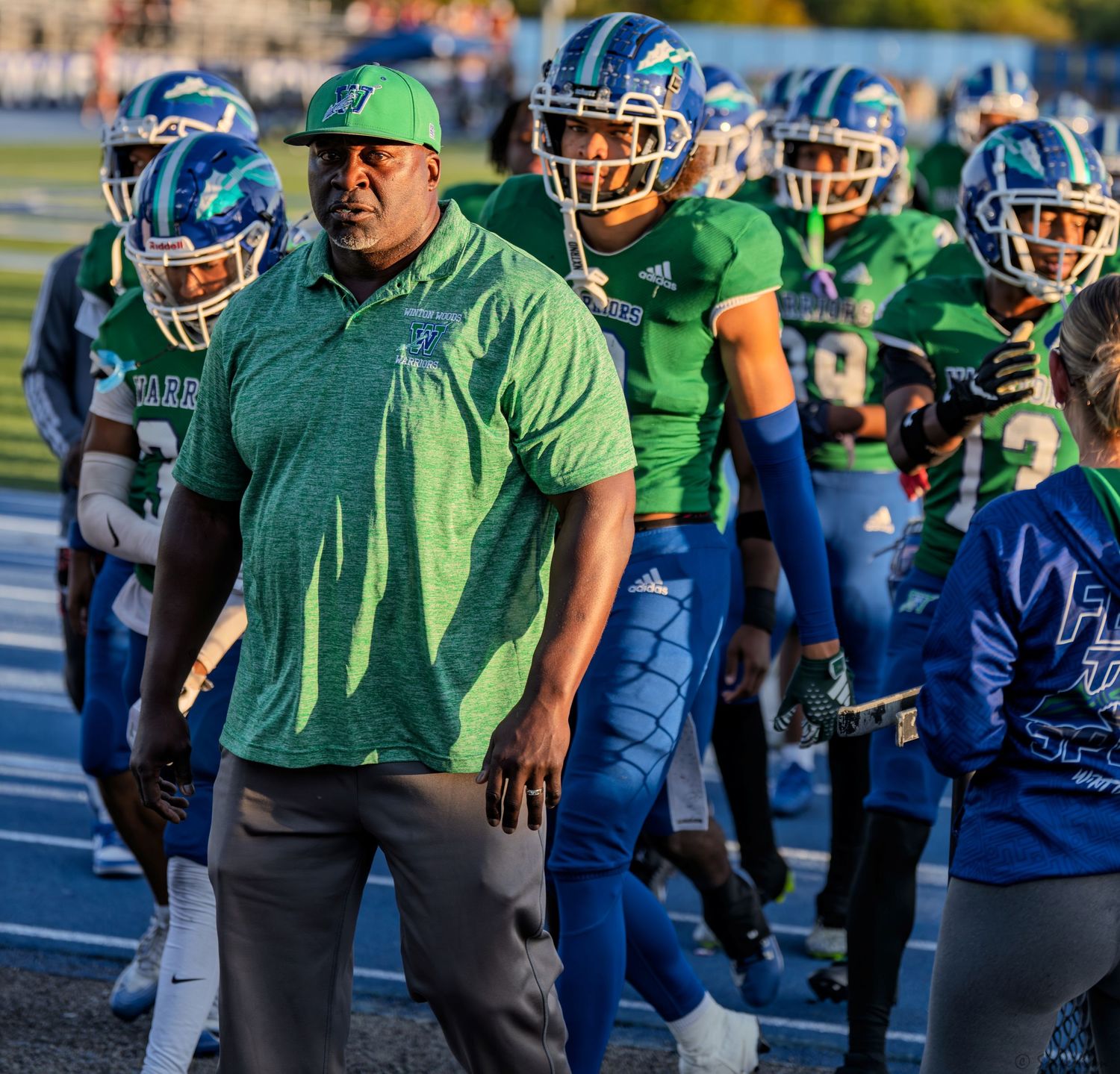 Football coach in green polo leads Warriors team in green and blue uniforms onto the field.