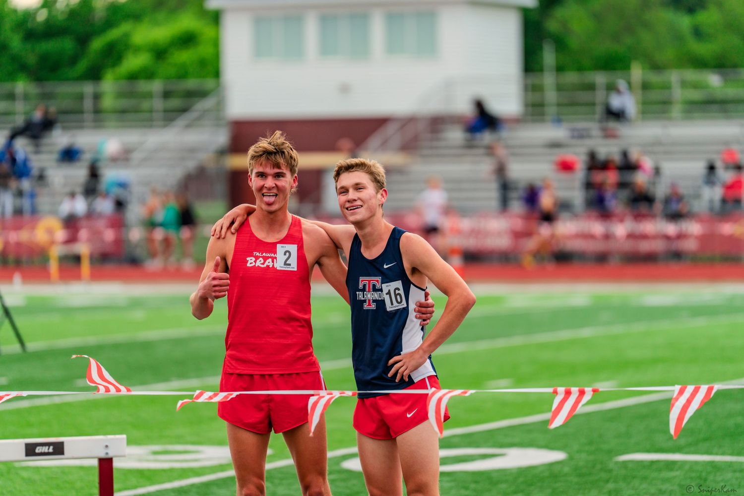 Two young male track runners pose together smiling near hurdles on a red athletic track.