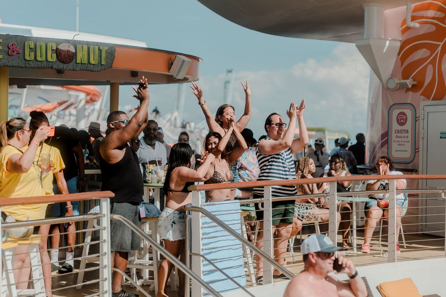 Excited cruise passengers dancing and celebrating on a sunny upper deck near a pool bar.