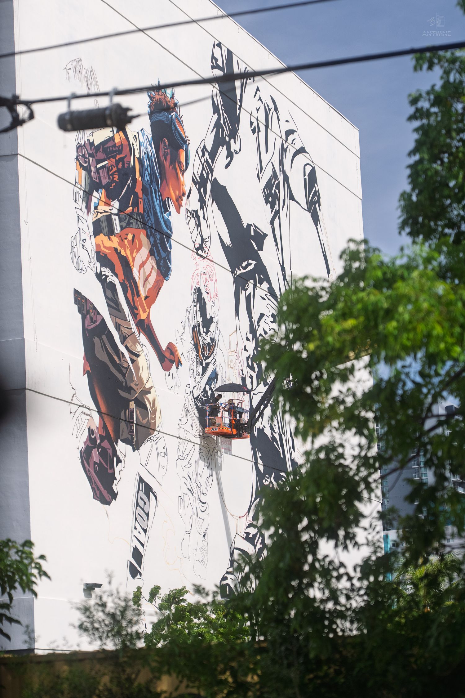 Artists on a crane painting a large colorful street art mural on a white building exterior wall.