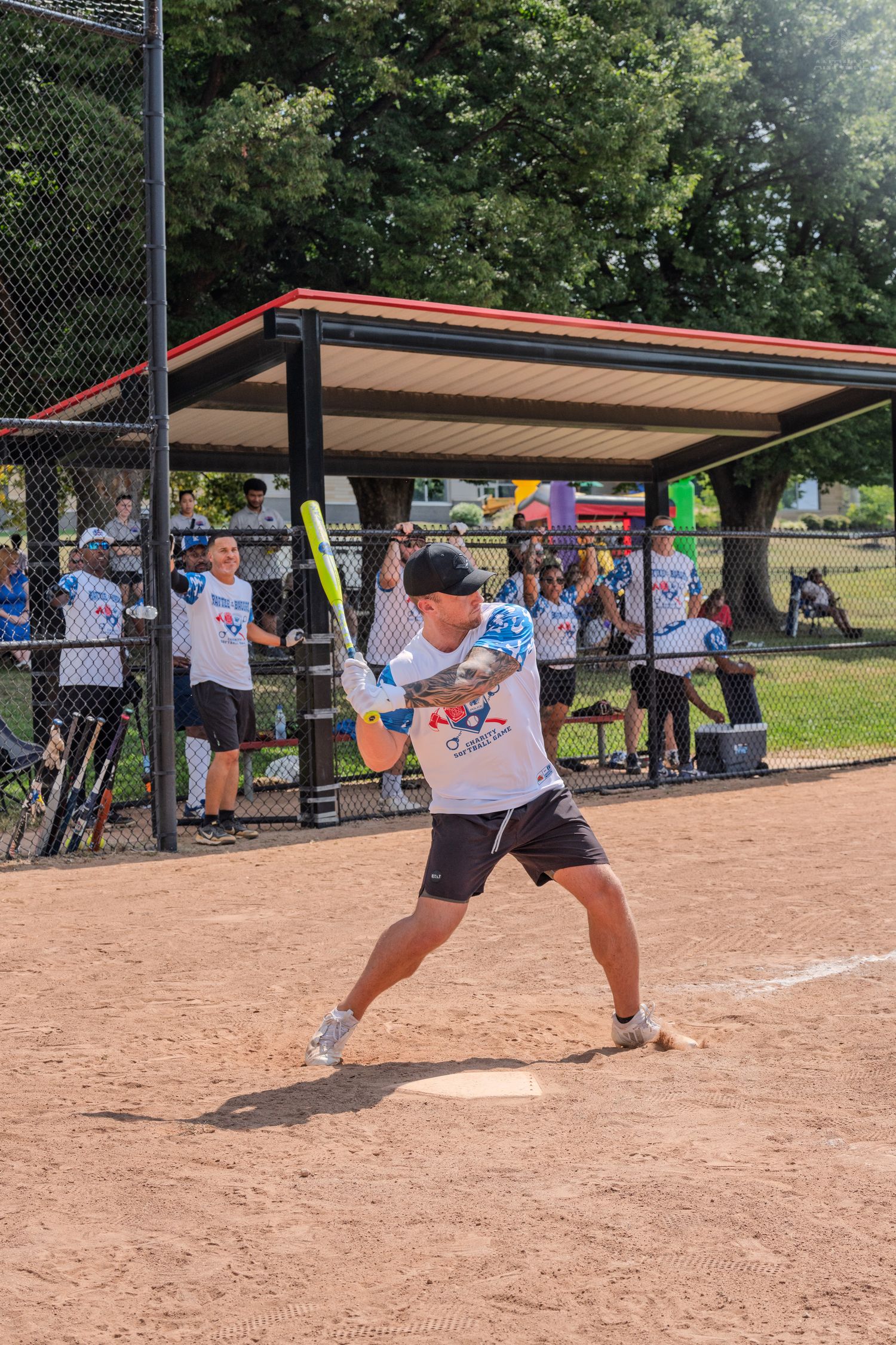 Batter in white jersey swinging yellow bat at home plate during charity softball game.