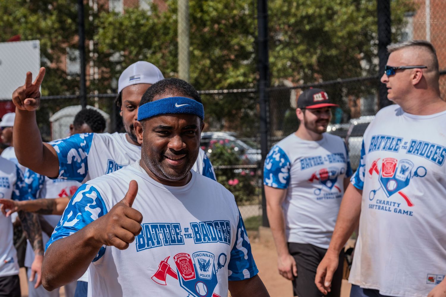 Excited man in Battle of the Badges charity softball shirt gives thumbs up while celebrating outdoors.