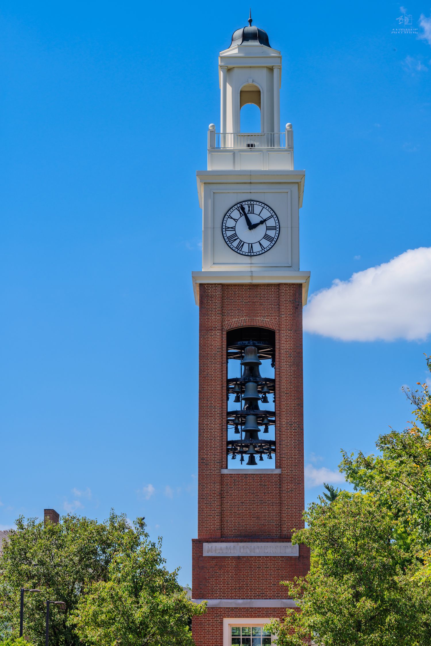 Tall red brick clock tower with white belfry, Roman numeral clock face, and multiple bells against blue sky.