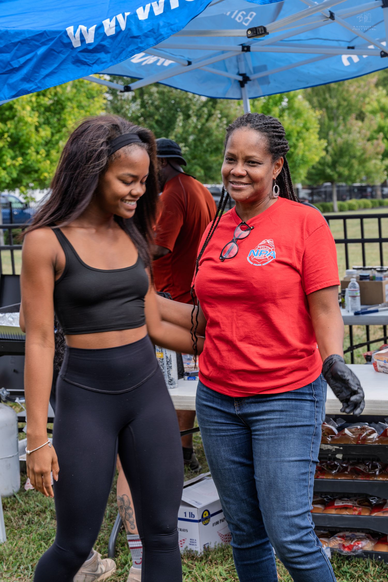 Two women smile together near food table under blue canopy at outdoor event.