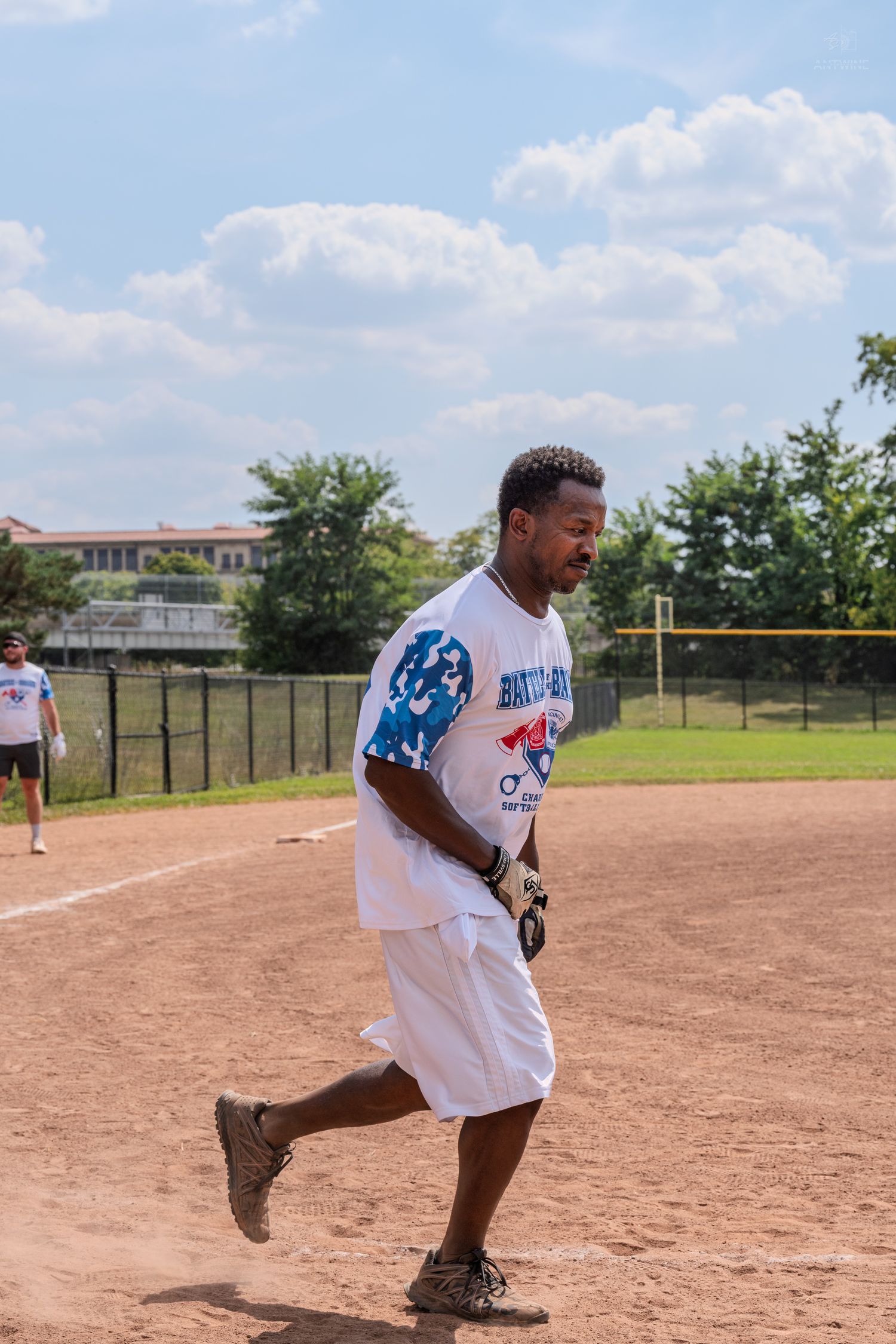 African American male softball player runs bases in white jersey on sunny dirt baseball field.