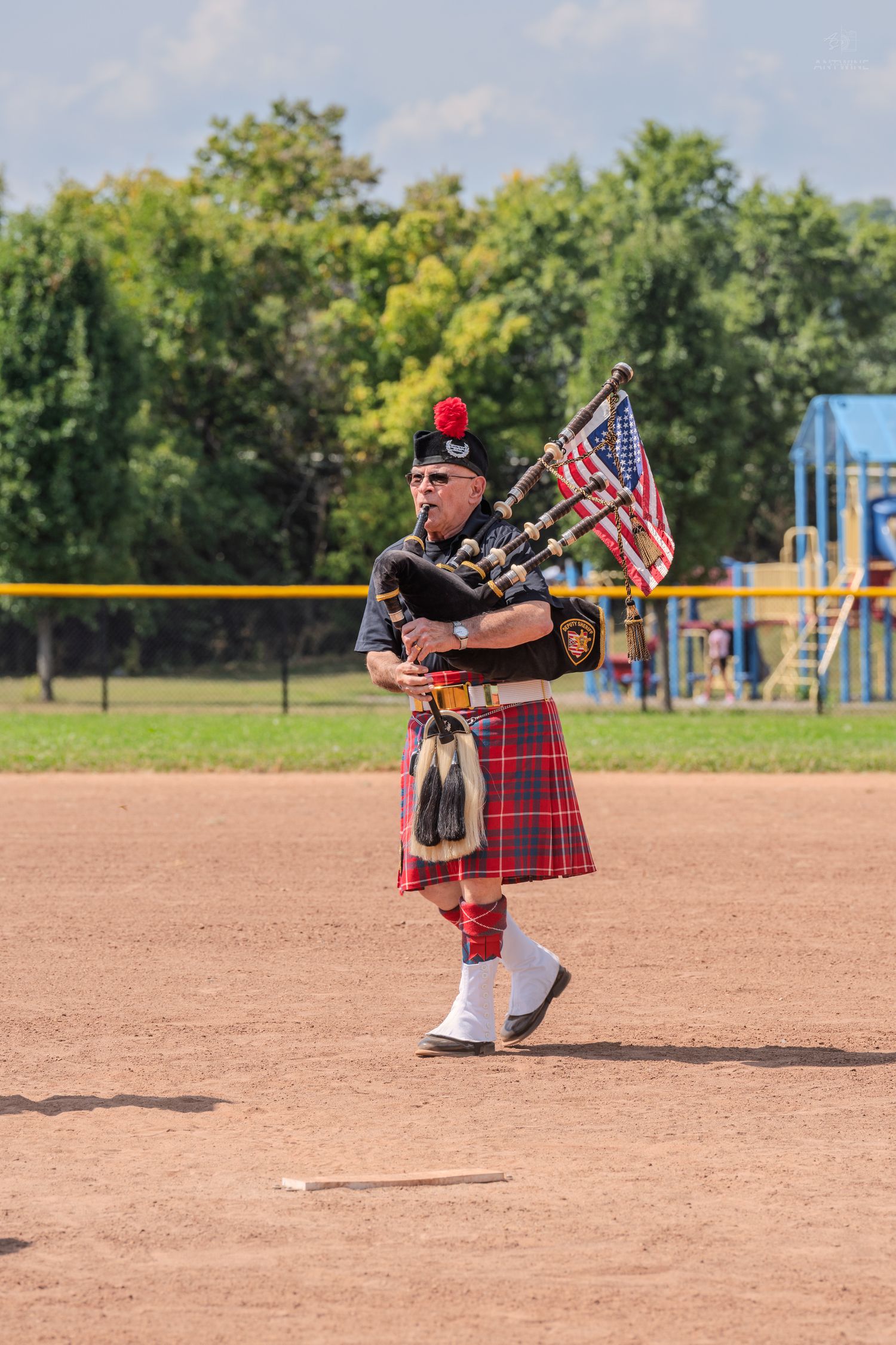 Bagpiper in red tartan kilt playing bagpipes with American flags on a baseball field outdoors.