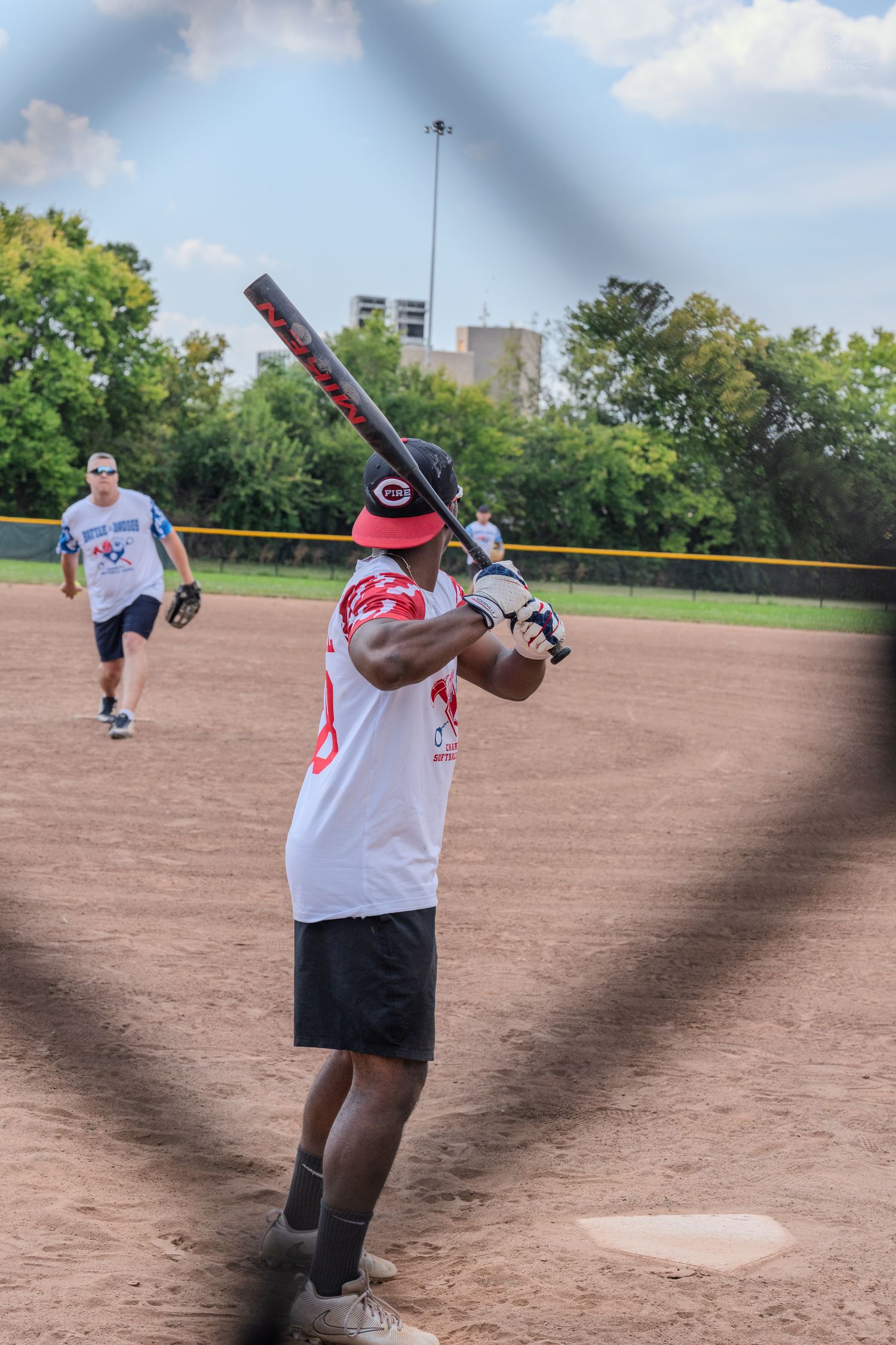 A softball batter in a white jersey and red cap stands ready at home plate.