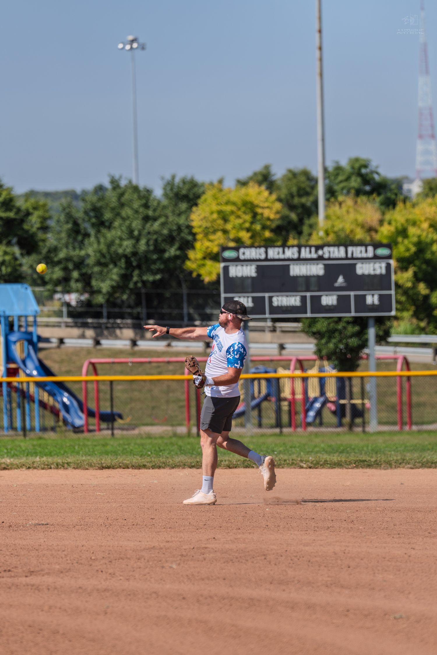 Female softball player throwing ball on dirt infield at Chris Helms All-Star Field on a sunny day.