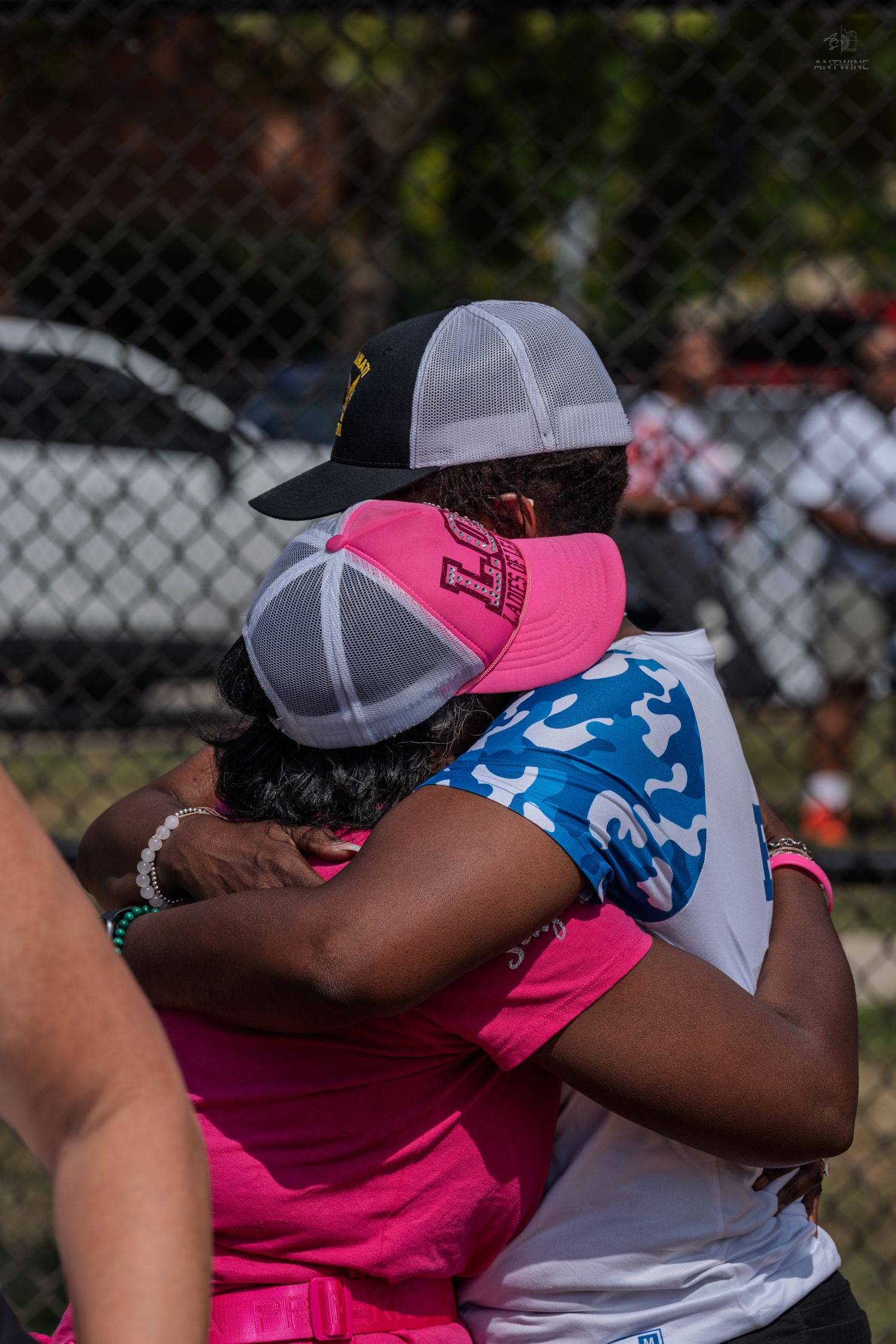 Two women sharing a warm hug outdoors, wearing colorful caps and casual attire at a sunny community event.