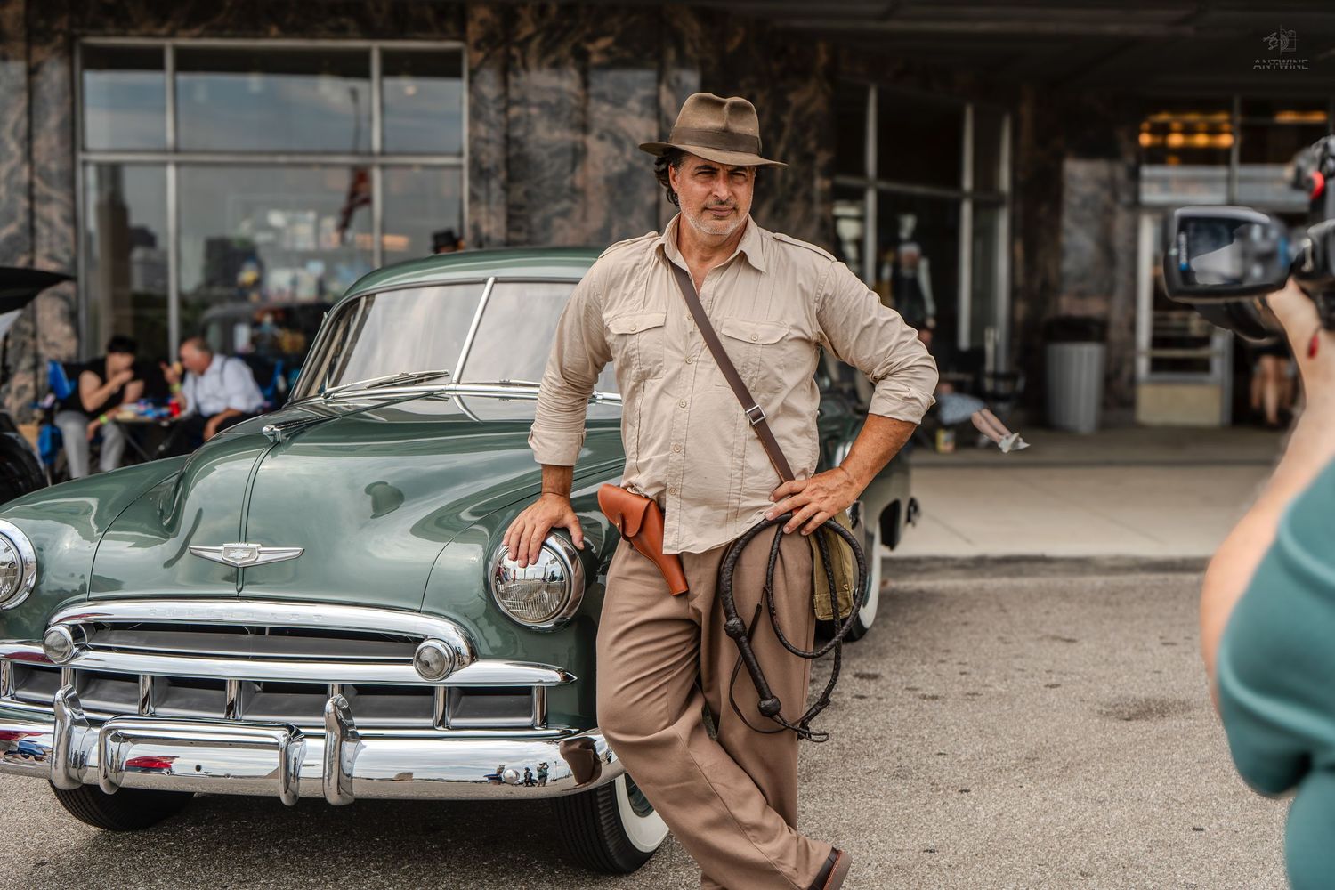 Man dressed as Indiana Jones leaning on a vintage green Chevrolet car outdoors.