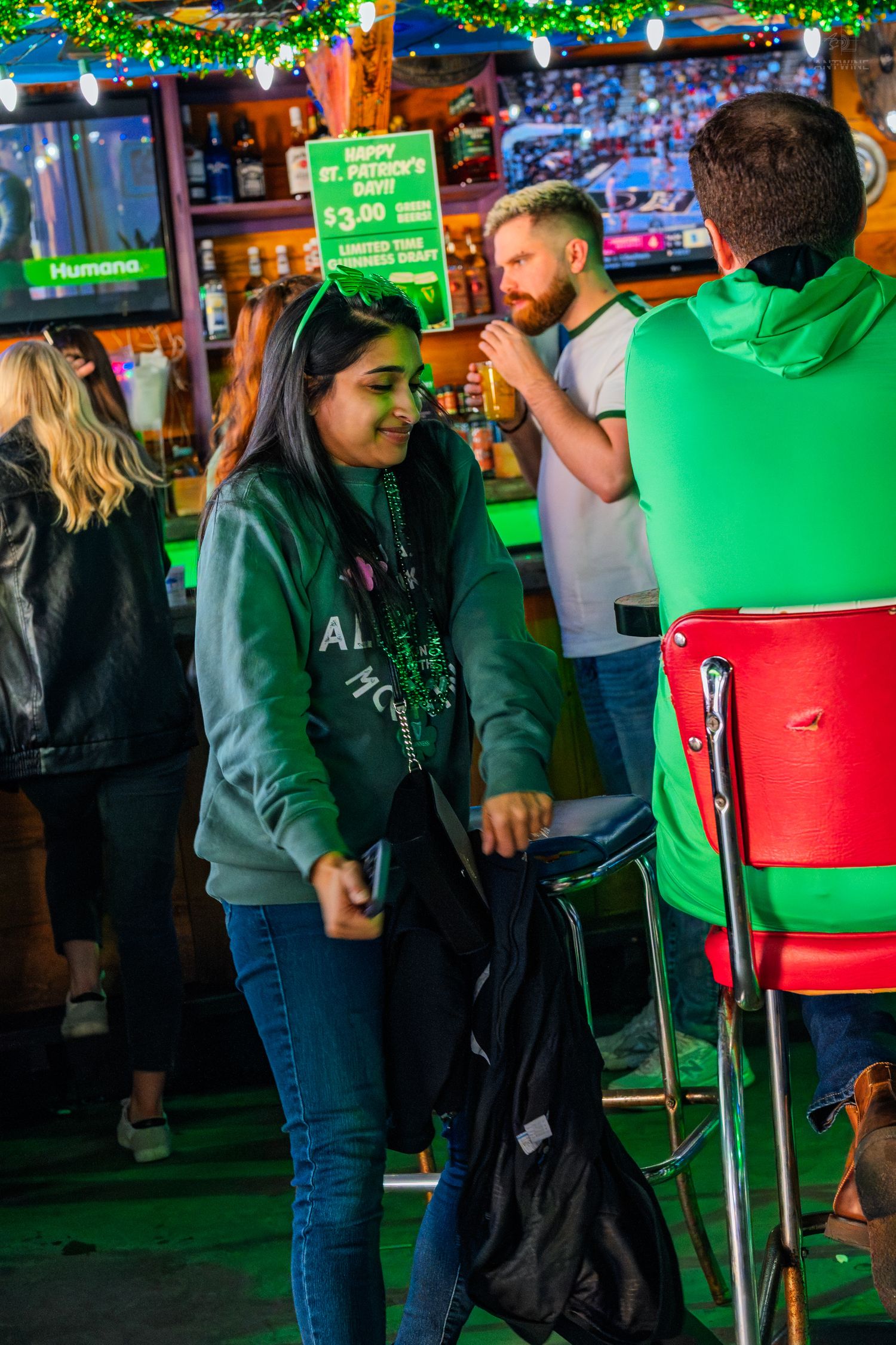 Woman in green hoodie with St. Patricks Day headband socializing at a festive bar celebration.