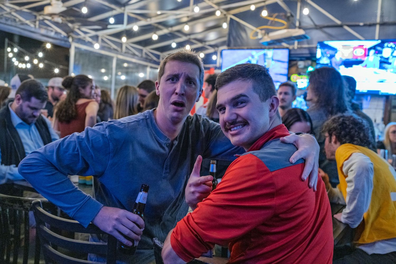 Two men smiling and posing together at a lively bar, one holding a beer bottle.