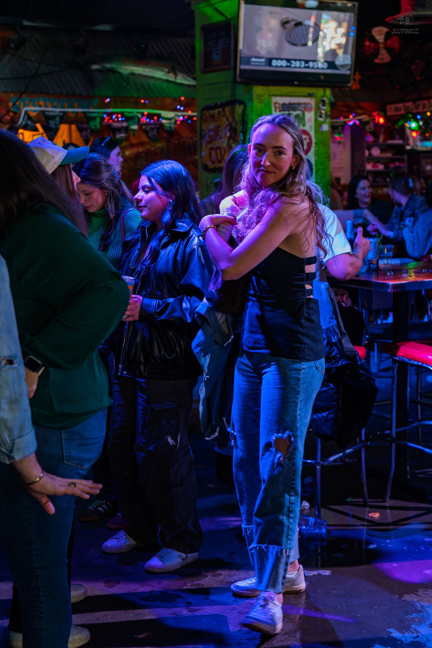 Young woman dancing in a vibrant neon-lit bar with friends gathered around her on the dance floor.
