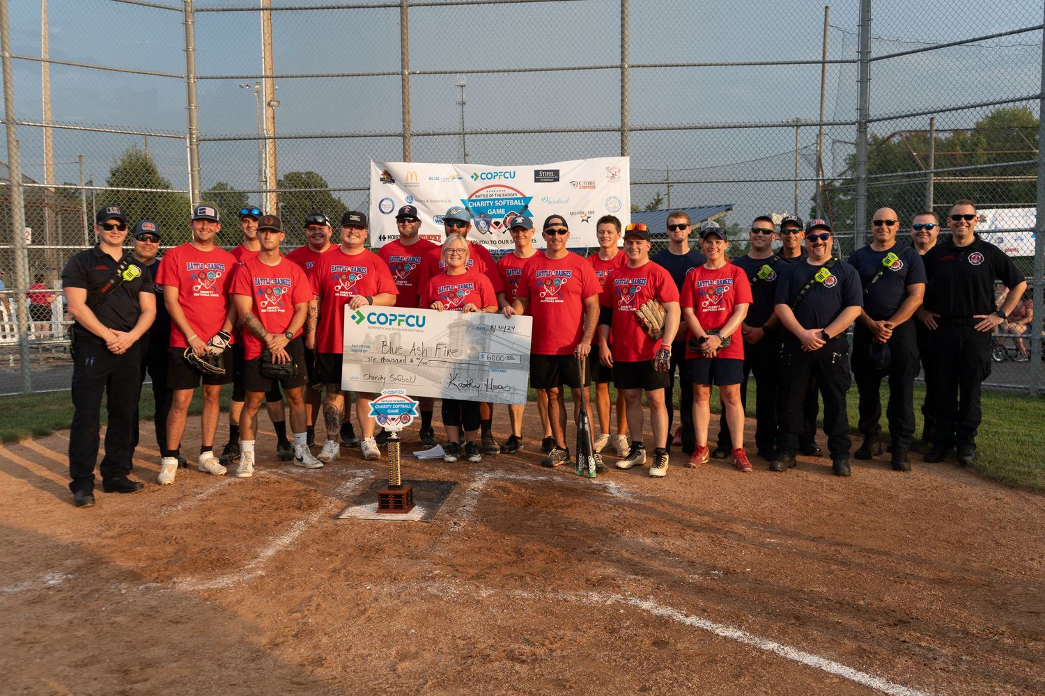 Group of firefighters and officials pose with oversized charity check at softball field event.