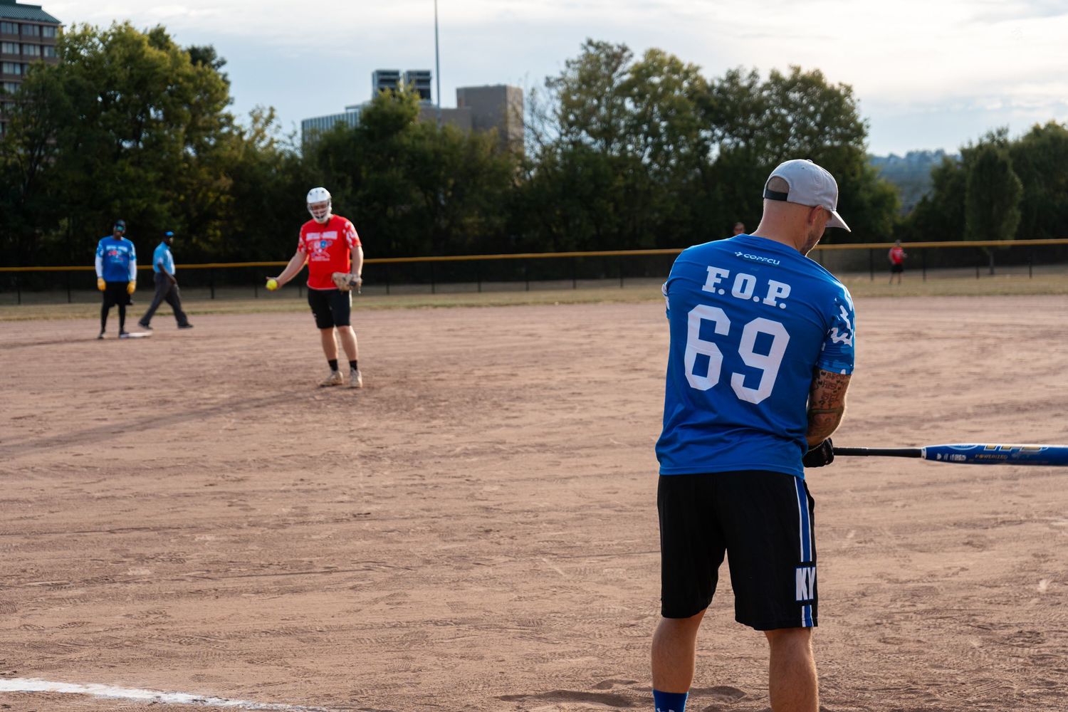 Player wearing FOP 69 blue jersey holds a bat during an outdoor softball game on a dirt field.