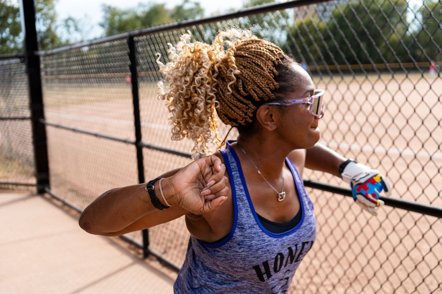Woman with curly blonde braids wearing a blue sleeveless shirt plays softball near a chain-link fence.