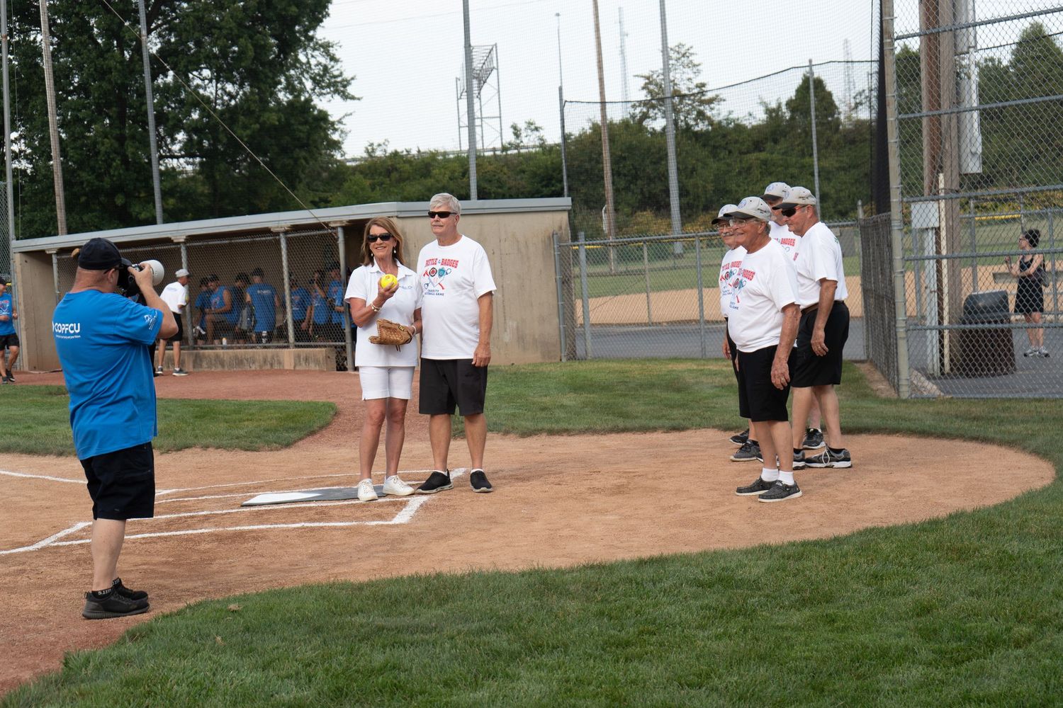 Group of people standing on a baseball diamond at an outdoor softball field during a sunny day event.