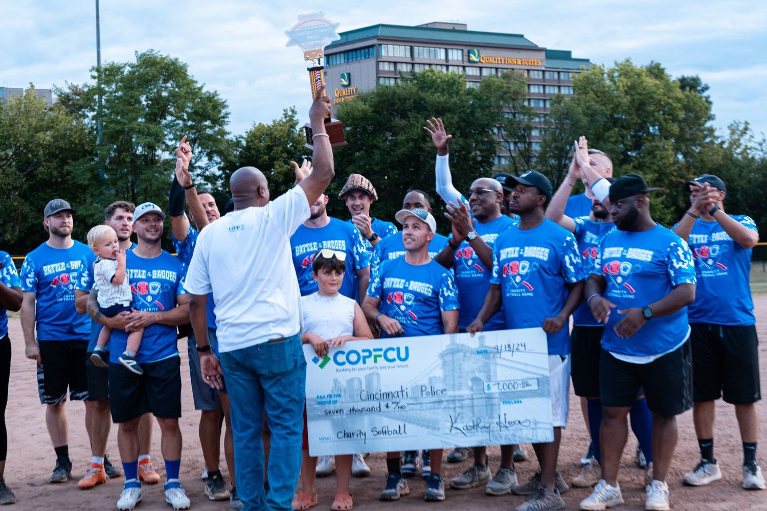 Winning softball team celebrates with a large COPFCU charity check at an outdoor baseball field.