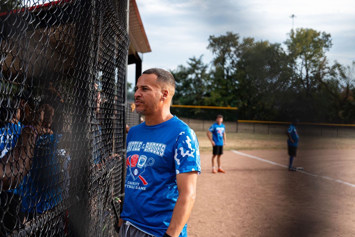 A man in a blue softball shirt leans against a chain-link fence watching players on a dirt field.