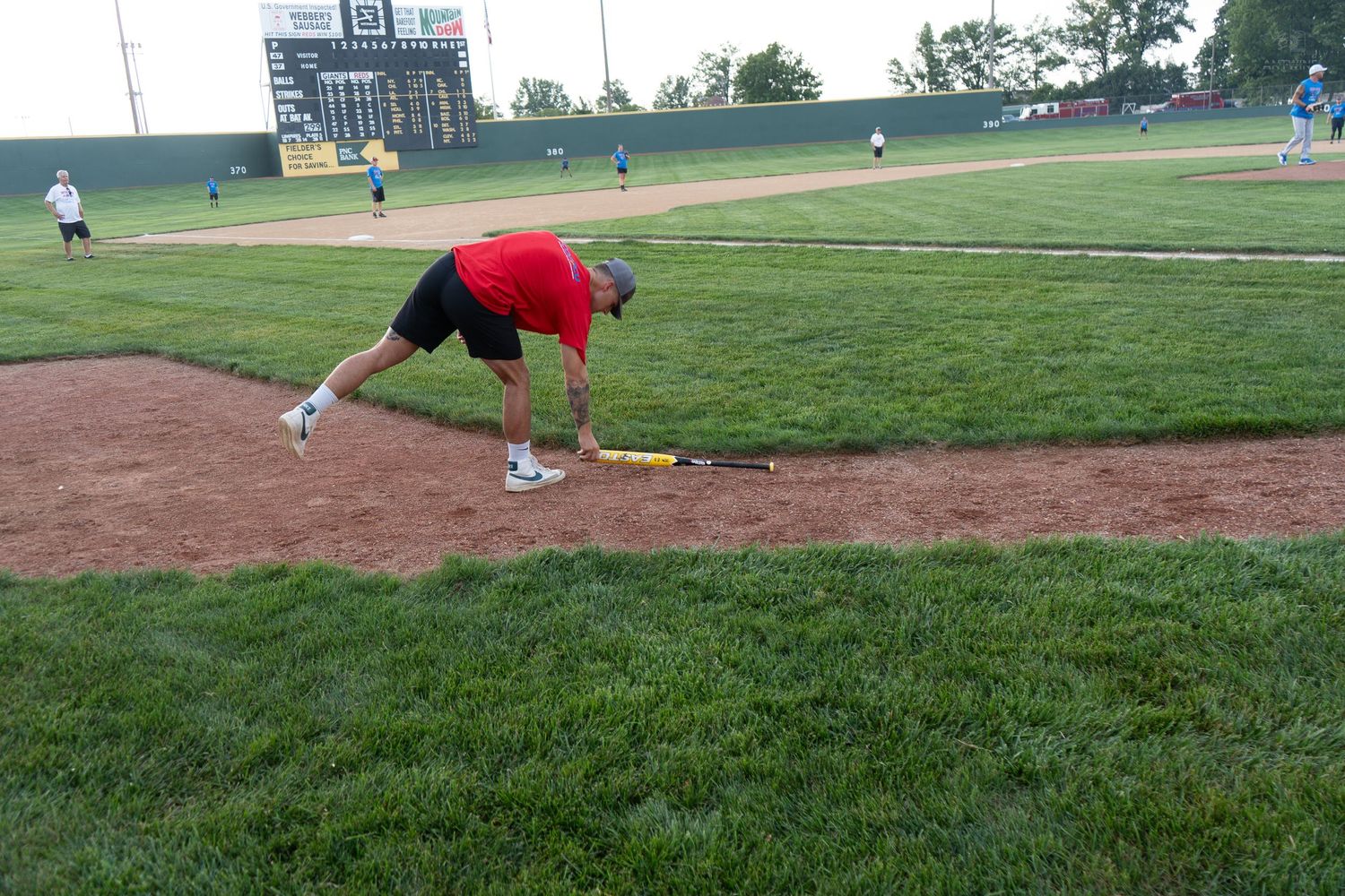 A man in a red shirt bending down to pick up a yellow bat on a baseball field pitcher's mound.
