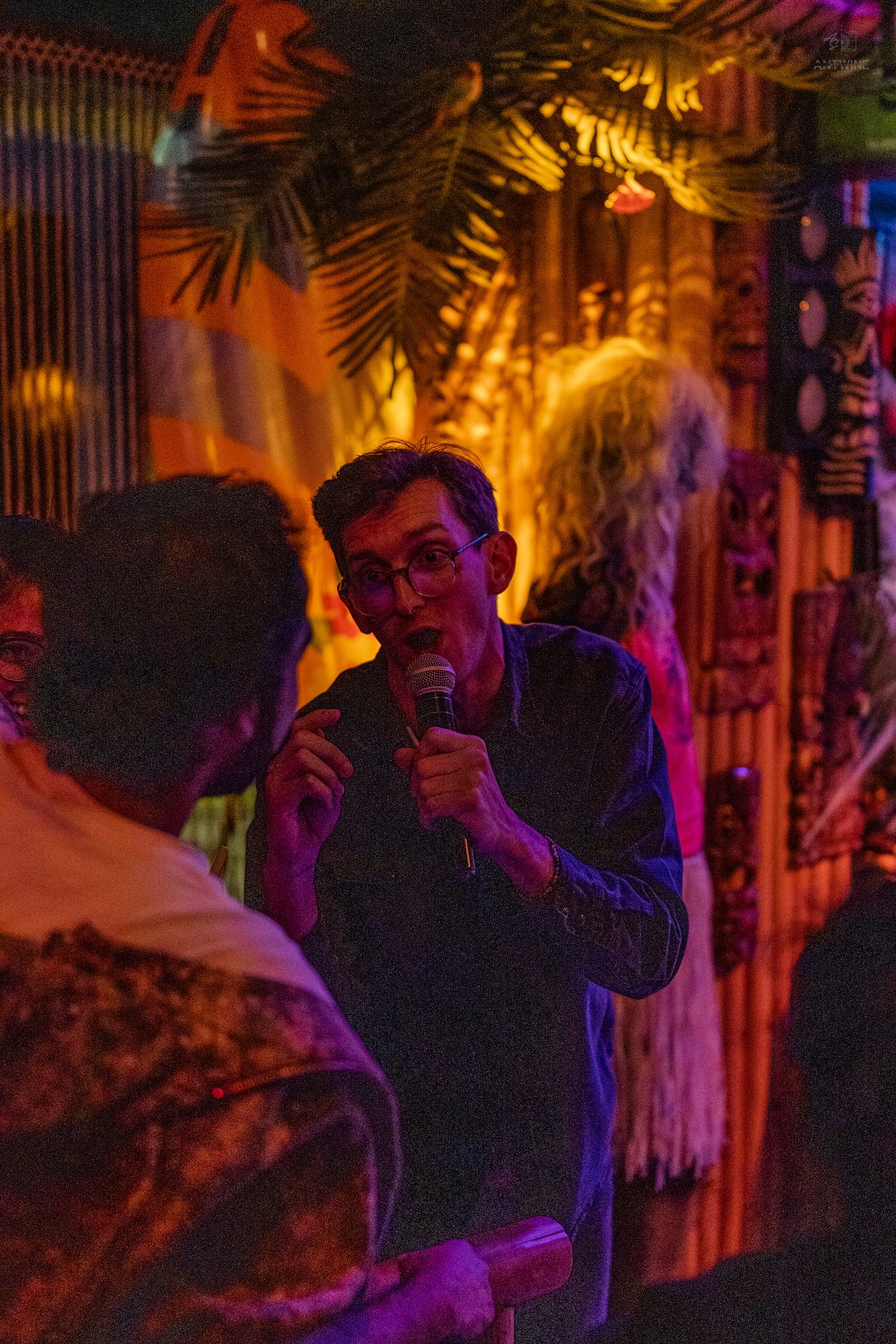Man singing karaoke with microphone at a tropical tiki bar with warm amber lighting and palm decor.