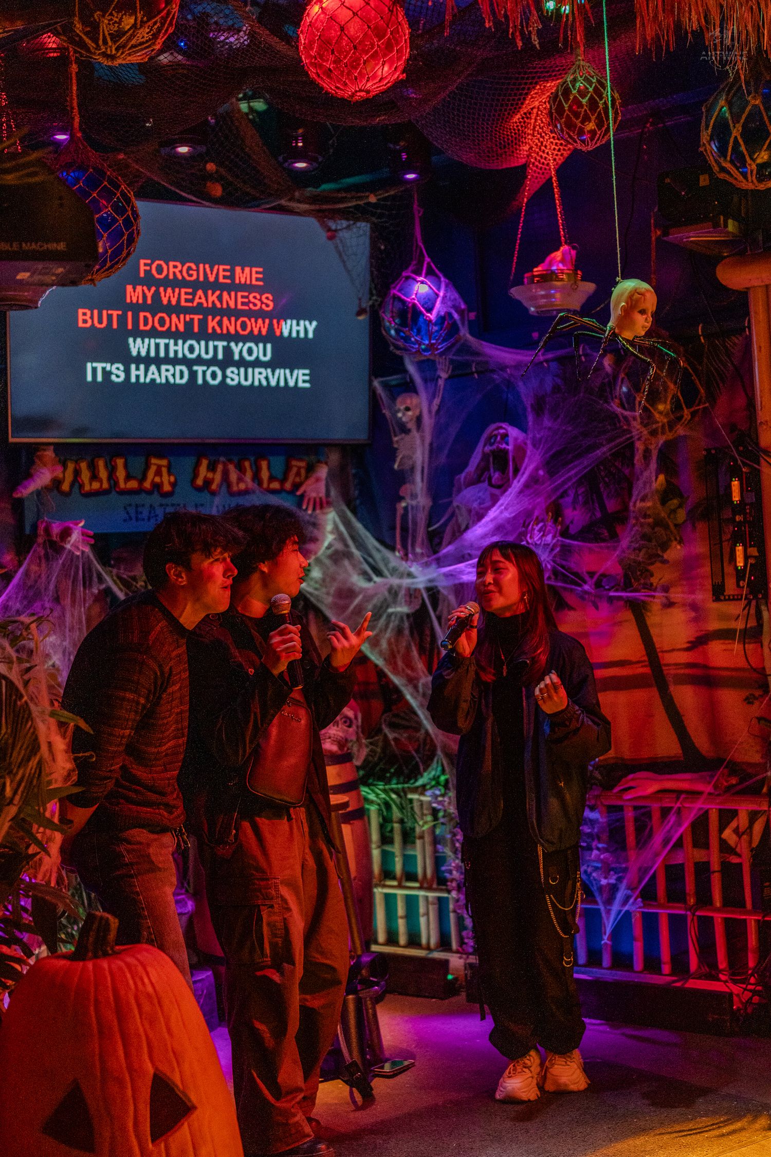 Two people singing karaoke duet at a Halloween-themed bar with spiderwebs, skeletons, and colorful neon lights.