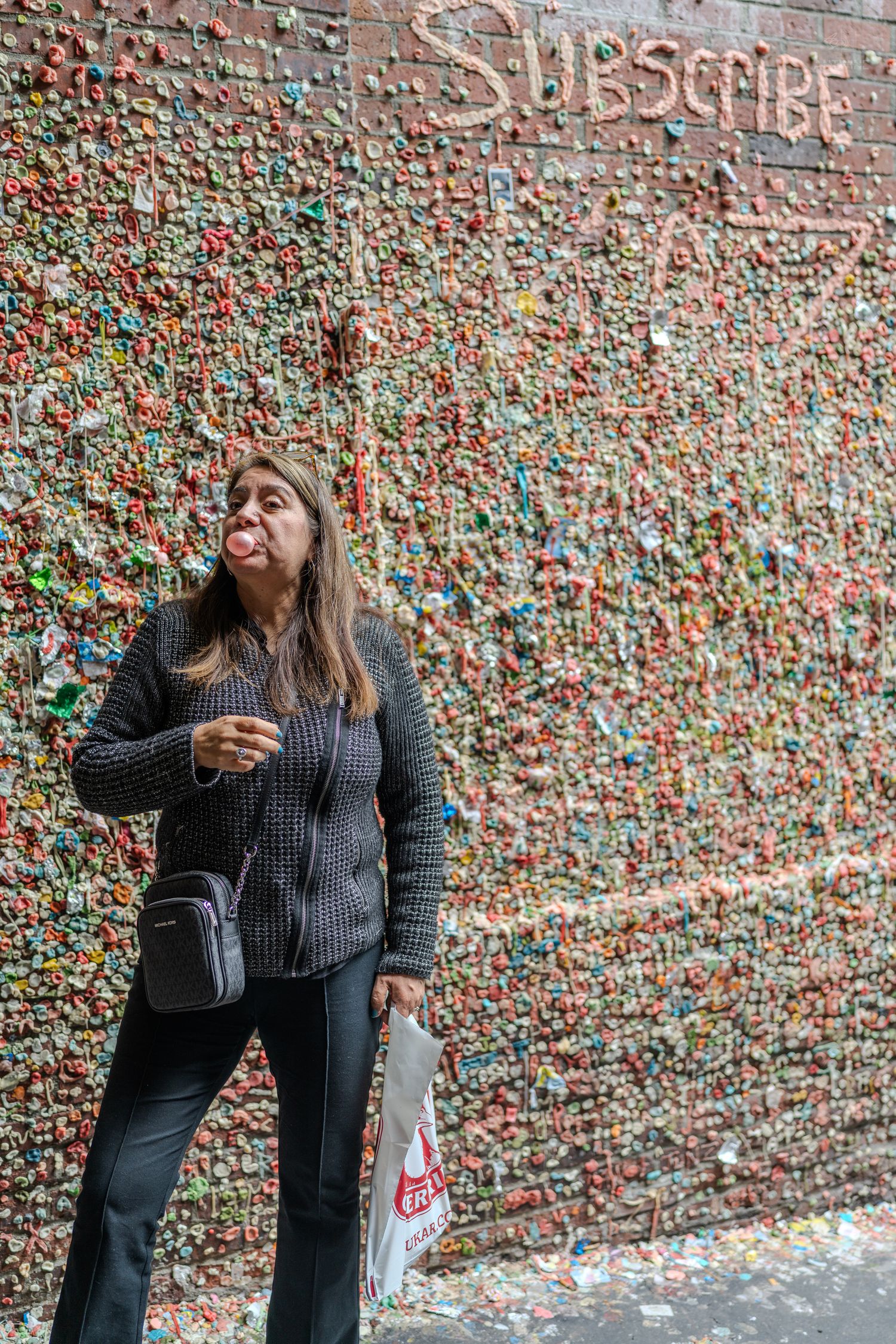 Woman blowing bubble gum bubble in front of Seattle's famous Gum Wall covered in colorful chewed gum.
